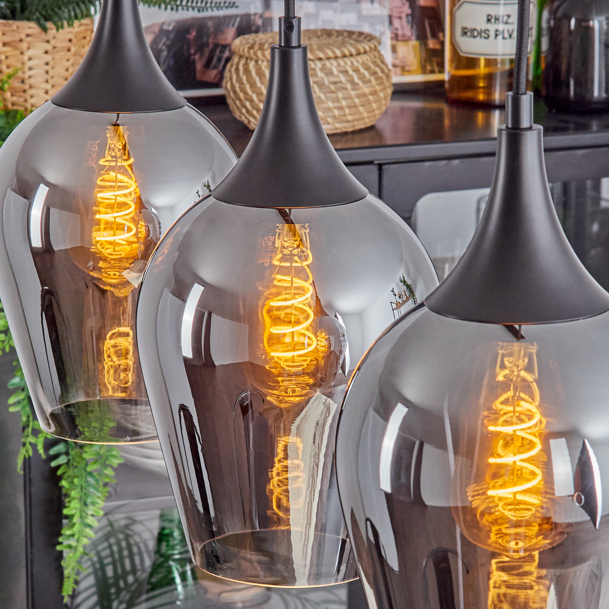 Drei Pendelleuchten mit Rauchglasschirmen und Wendelglühlampen. Im Hintergrund Körbe und Grünpflanzen.