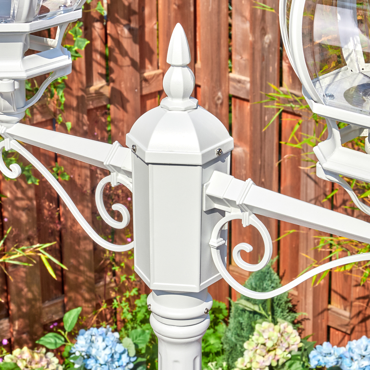 Weißer, verzierter Laternenpfahl mit Holzzaun im Hintergrund und bunten Blumen. Der Fokus liegt auf dem detaillierten Design.