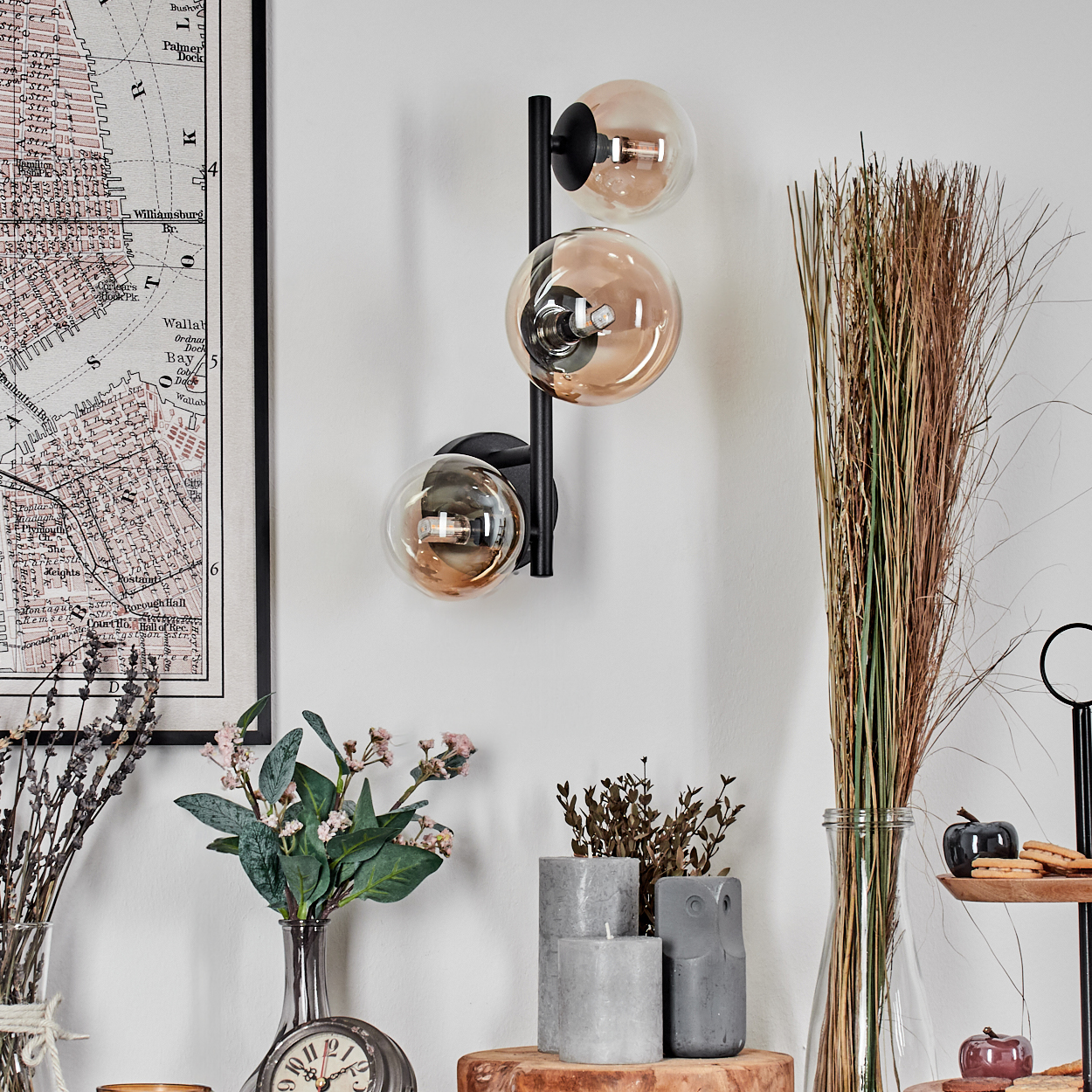 Eine Wand mit Karte, moderner Lampe, Blumen, Kerzen und Dekoartikeln. Die Lampe hat drei runde Glasschirme.