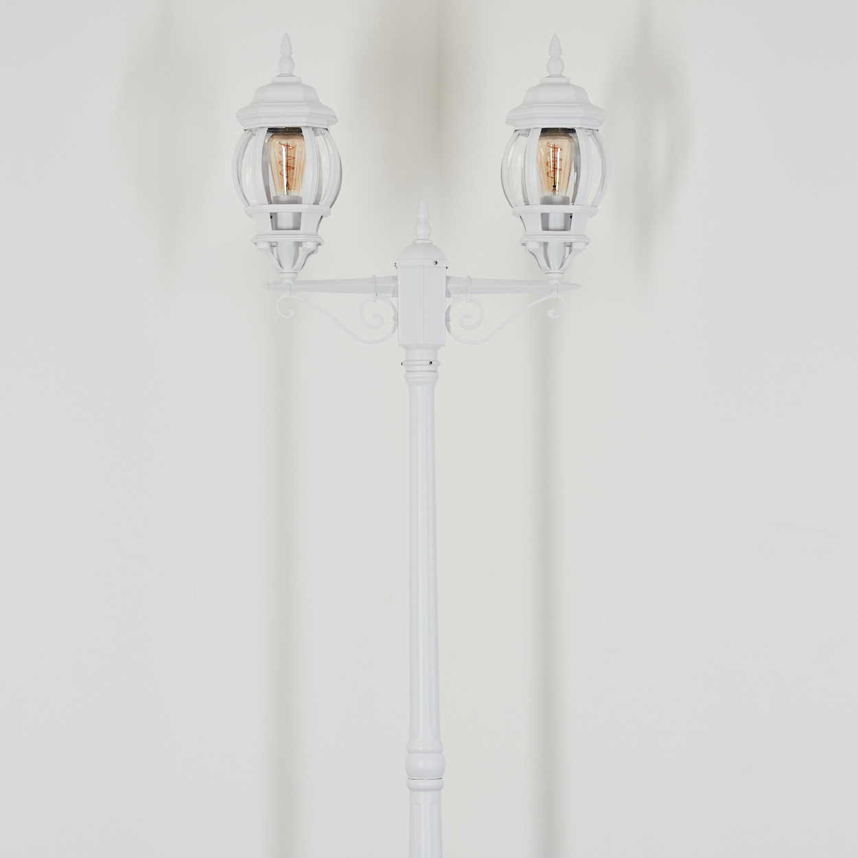 Eine weiße dekorative Straßenlaterne mit zwei Lampen. Jede Lampe hat eine sichtbare Glühbirne. Der Hintergrund ist eine weiße Wand.