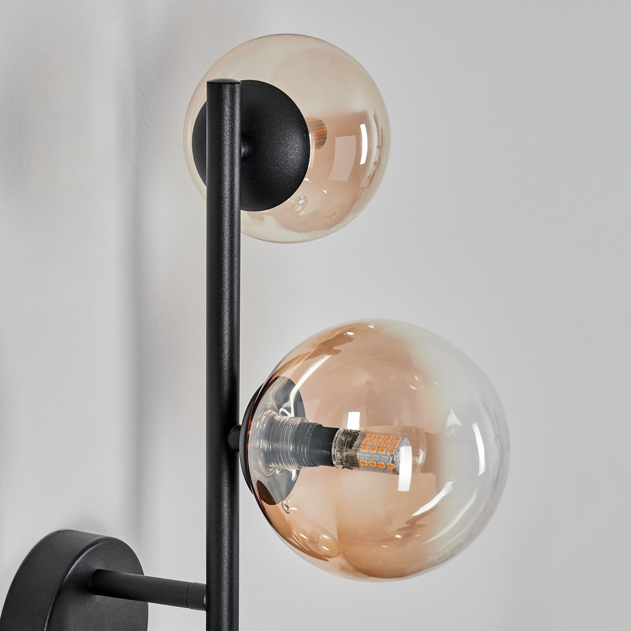 Eine moderne Wandlampe mit zwei kugelförmigen, bernsteinfarbenen Glasschirmen. Ein schwarzer Metallrahmen hält die Kugeln an einer weißen Wand.