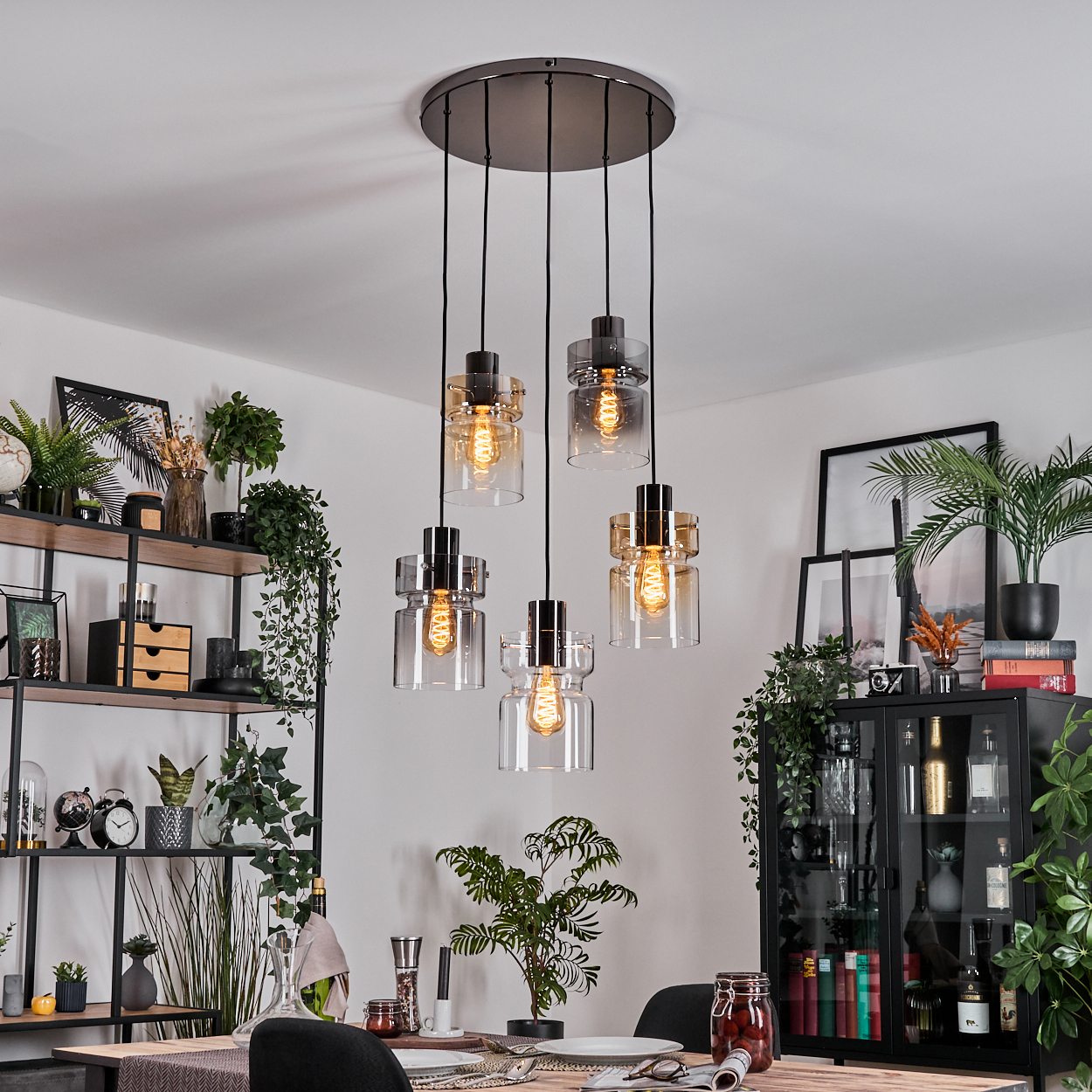Esszimmer mit Hängelampen, Pflanzen und einem schwarzen Schrank. Tisch mit Gläsern und Tellern gedeckt.