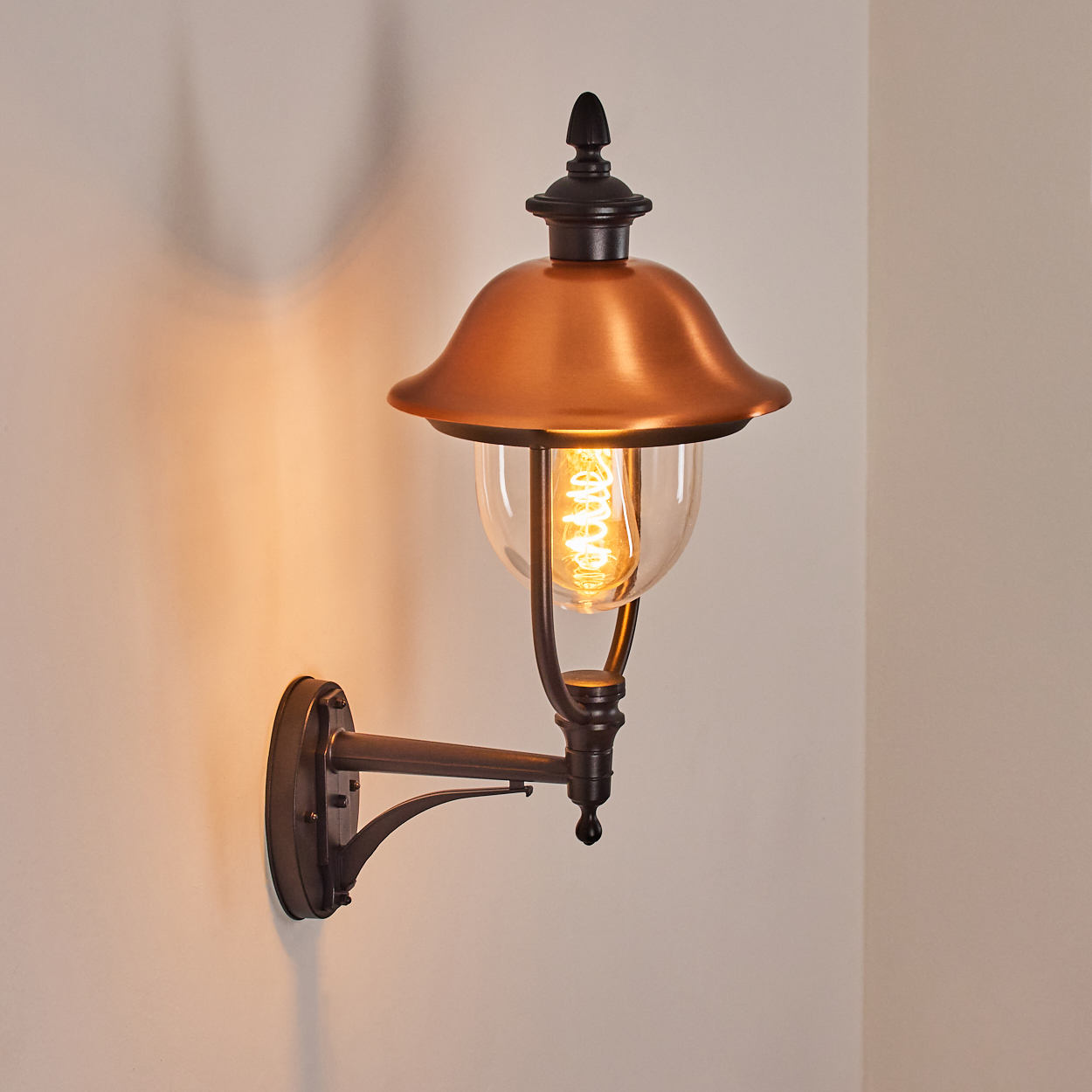 Eine an der Wand befestigte Kupfer- und schwarze Außenlampe mit einem klaren Glaskolben und einer sichtbaren Glühbirne. Die Wand ist hellbeige.