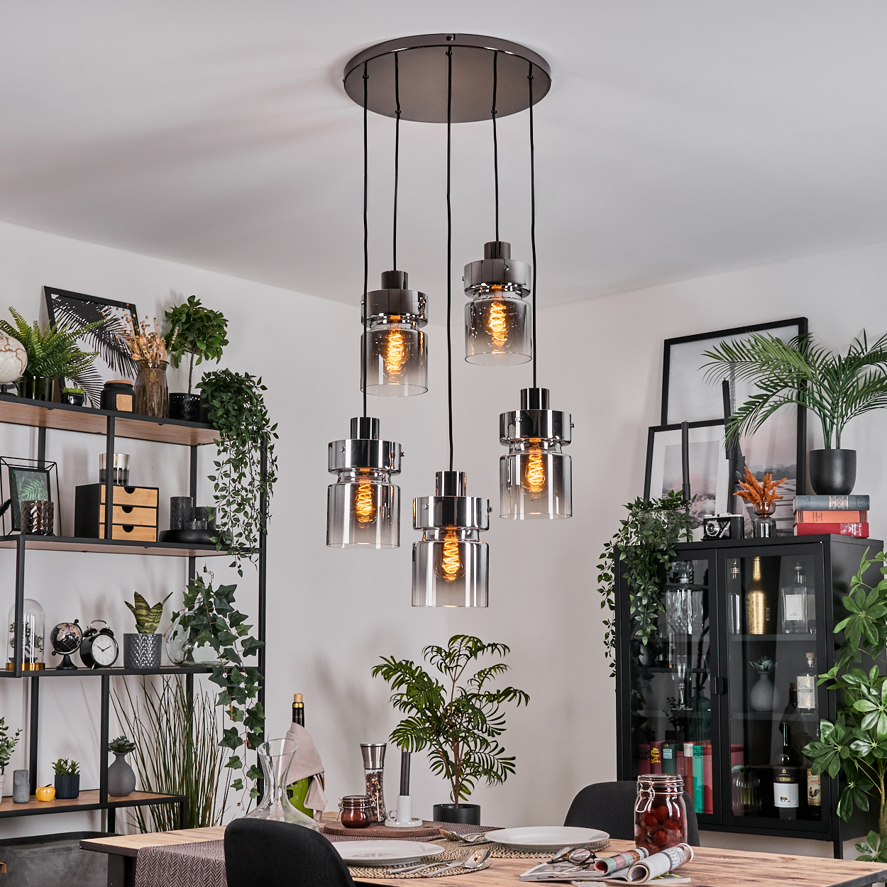 Esszimmer mit Hängelampen, Pflanzen und einem gedeckten Holztisch. Regale und ein Schrank sind im Hintergrund zu sehen.