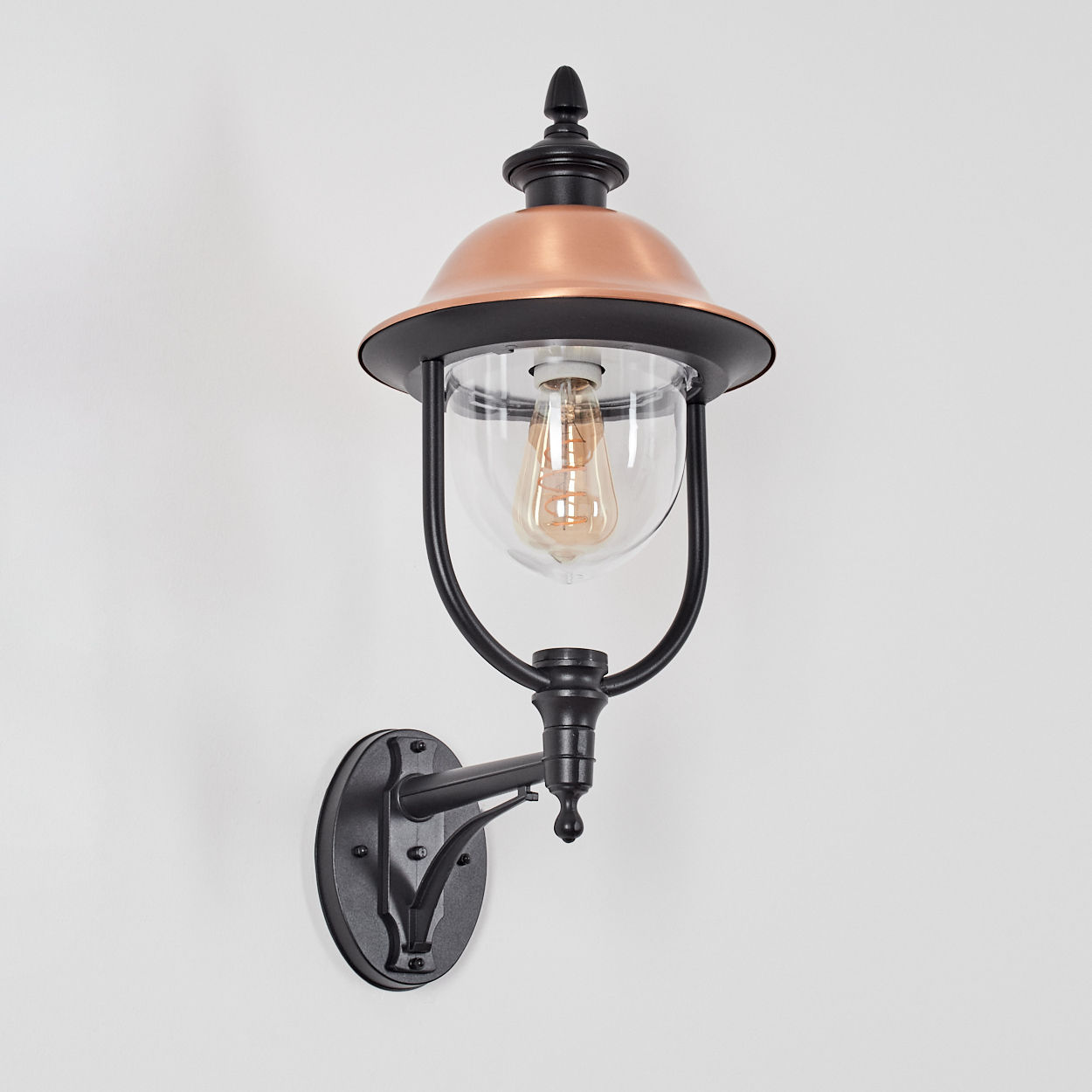 Eine an der Wand befestigte Außenlampe. Sie hat einen kupferfarbenen Deckel, einen schwarzen Rahmen und klares Glas, das eine Glühbirne umgibt. An einer weißen Wand montiert.