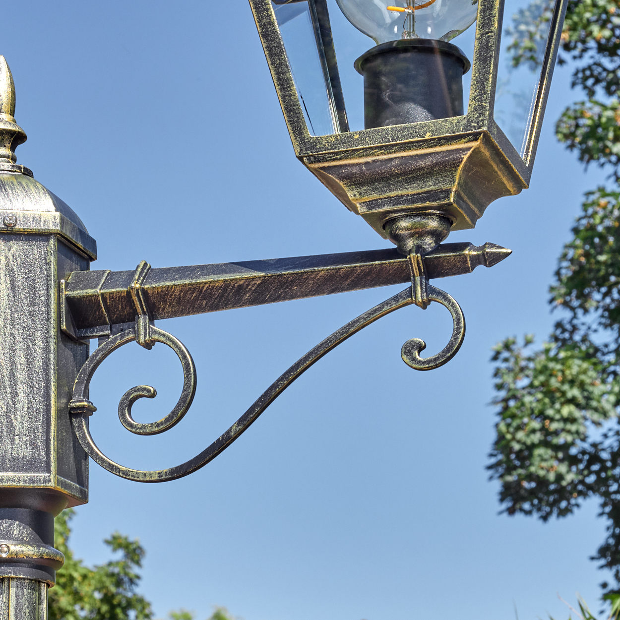 Eine verzierte Straßenlaterne im antiken Stil. Sie hat ein Glasgehäuse und eine dekorative Halterung vor blauem Himmel mit Bäumen.
