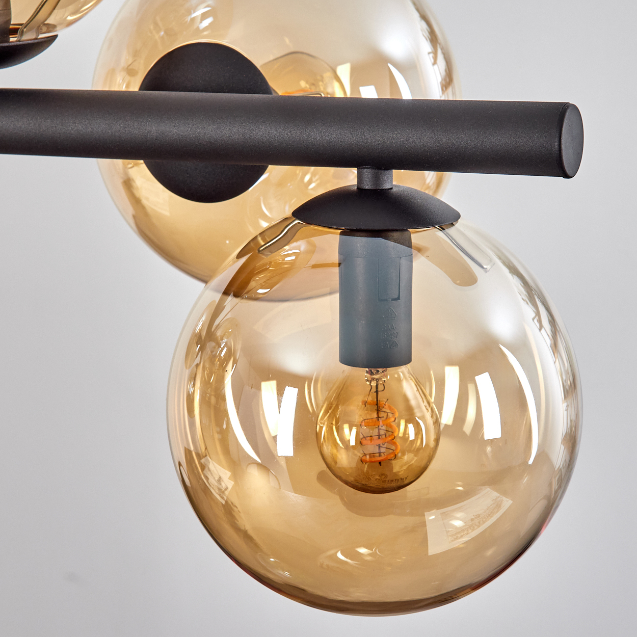 Nahaufnahme eines modernen Kronleuchters mit drei kugelförmigen, bernsteinfarbenen Glasschirmen, schwarzen Metallstangen und sichtbaren Glühbirnen.