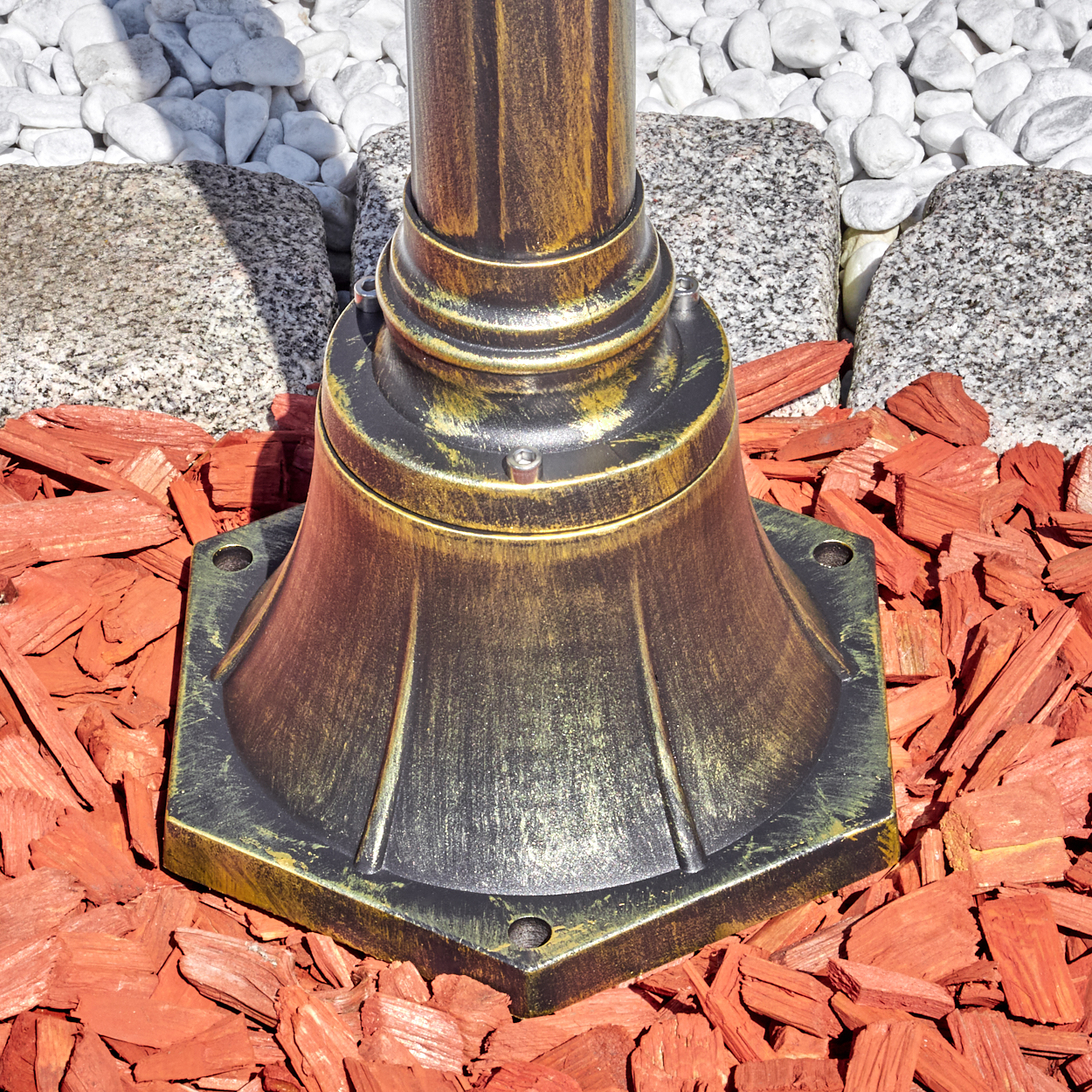 Ein bronzefarbener Lampensockel steht auf roter Mulch. Im Hintergrund befinden sich weiße Steine und Granitblöcke.