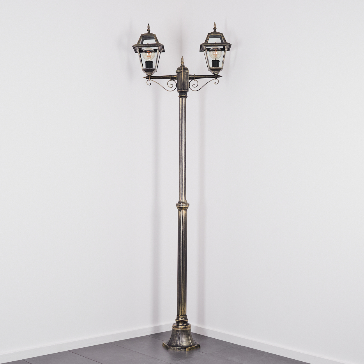 Eine verzierte, bronzefarbene Straßenlaterne mit zwei Lampen. Sie steht in einer Ecke vor einer weißen Wand und einem grauen Boden.