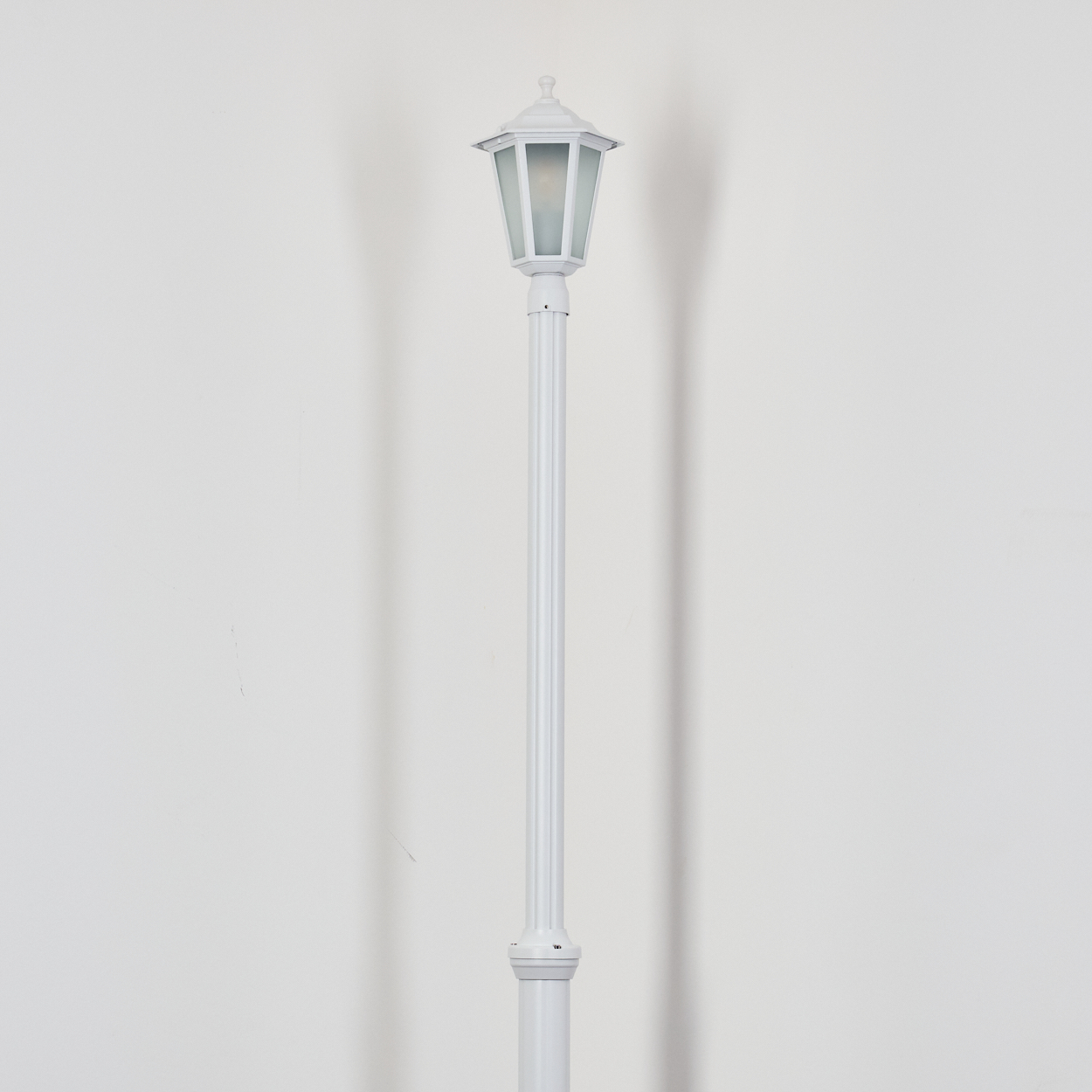 Eine weiße Straßenlaterne steht vor einer weißen Wand. Die Lampe hat einen Laternenaufsatz und eine lange Stange. Ein Schatten ist an der Wand.