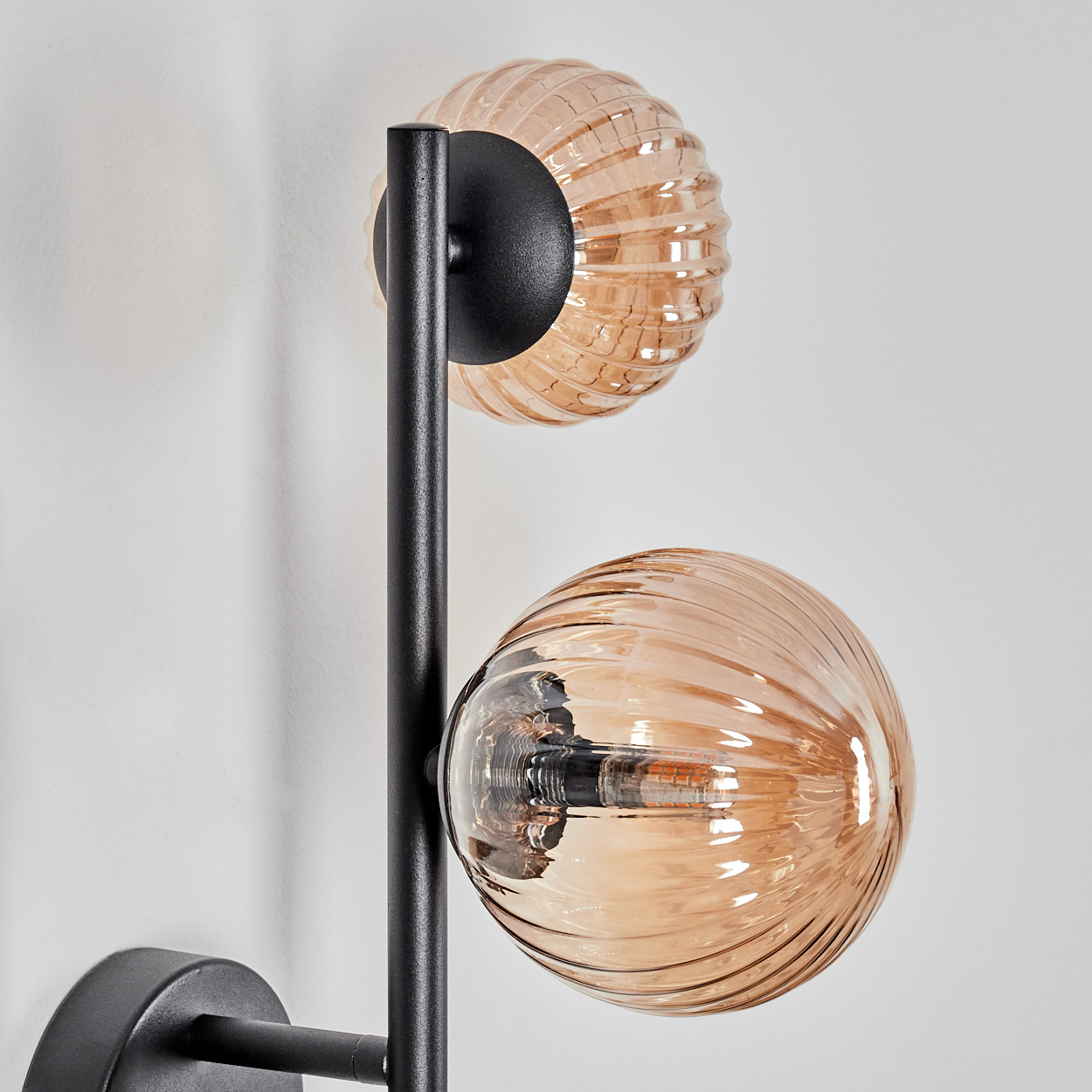 Eine moderne Wandlampe mit zwei bernsteinfarbenen Glaskugeln. Schwarzer Metallrahmen. Weißer Wandhintergrund.
