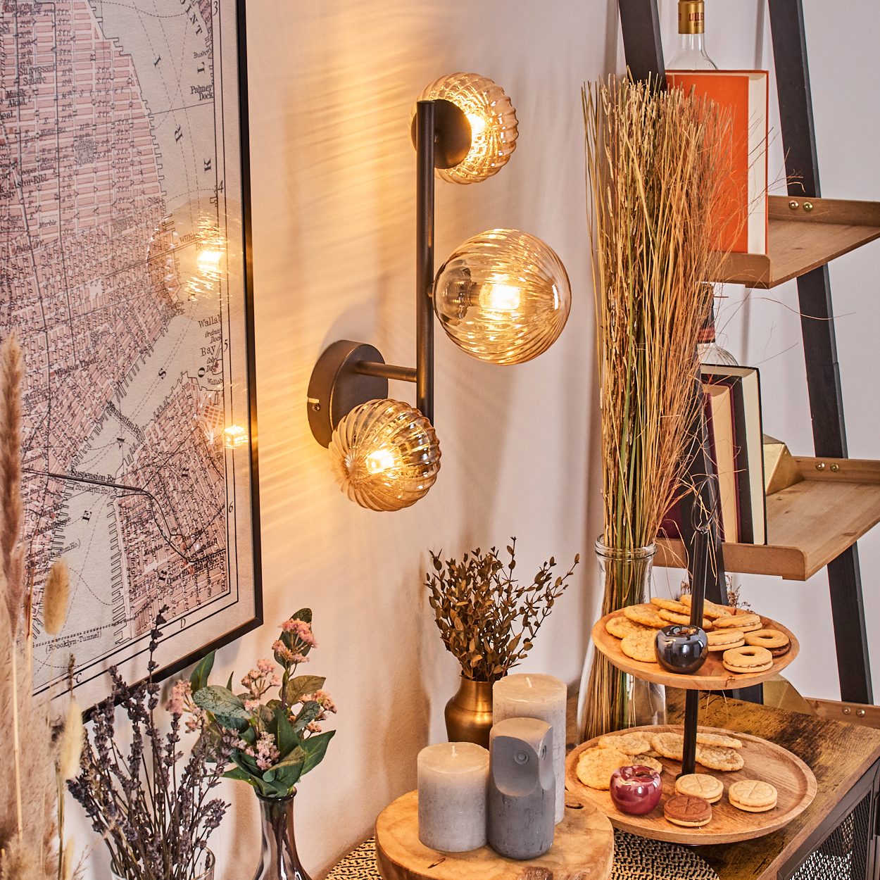 Eine dekorative Szene mit Wandlampe, Karte, Blumen, Kerzen und Keksen auf einem Holzregal. Warme Beleuchtung und gemütliche Atmosphäre.
