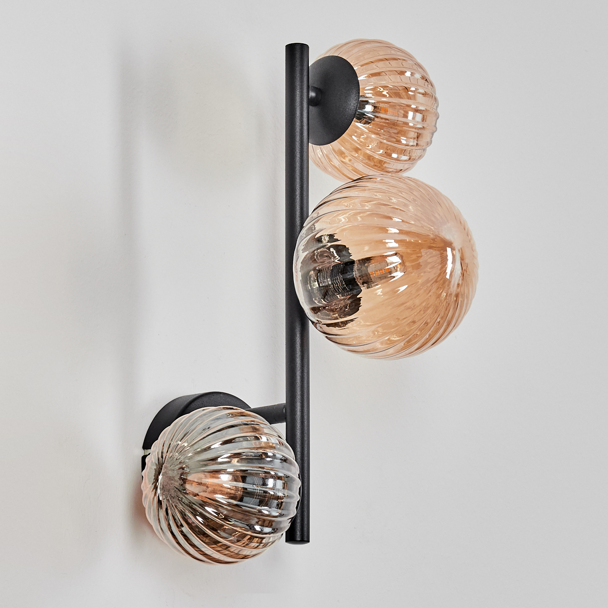 Eine moderne Wandlampe mit drei bernsteinfarbenen Glaskugelschirmen. Die Lampe hat einen schwarzen Metallrahmen und ist an einer weißen Wand befestigt.