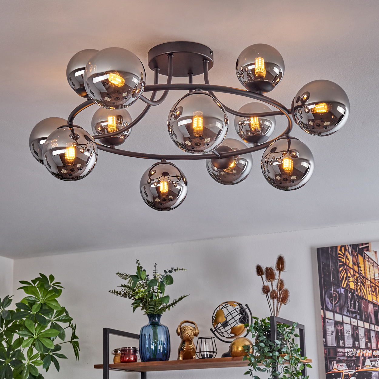 Eine moderne Deckenleuchte mit vielen kugelförmigen Glasschirmen. Darunter ein Regal mit Dekorationen wie Pflanzen, einem Globus und einem Gemälde.