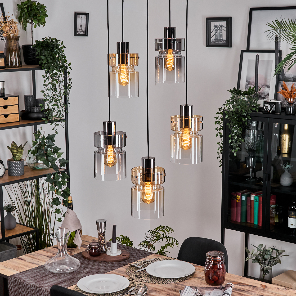 Esszimmer mit Hängelampen. Tisch mit Tellern, Besteck und Dekorationen. Pflanzen und Regale mit Gegenständen im Hintergrund.