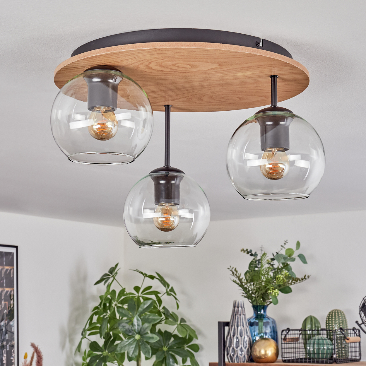 Eine moderne Deckenlampe mit drei Glaskugeln, Holzsockel und schwarzen Akzenten. Der Hintergrund umfasst Pflanzen und Dekoration.