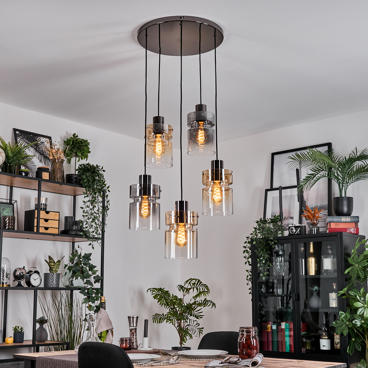 Esszimmer mit Hängelampen, Regalen und einem Tisch, der für eine Mahlzeit gedeckt ist. Pflanzen und Dekorationen sind sichtbar.