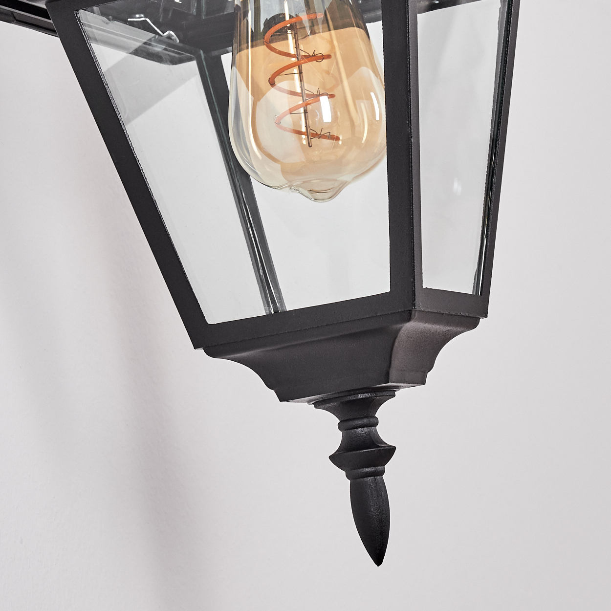 Nahaufnahme einer schwarzen Außenlampe im Vintage-Stil mit Glaspaneelen und einer leuchtenden Glühbirne vor einer weißen Wand.