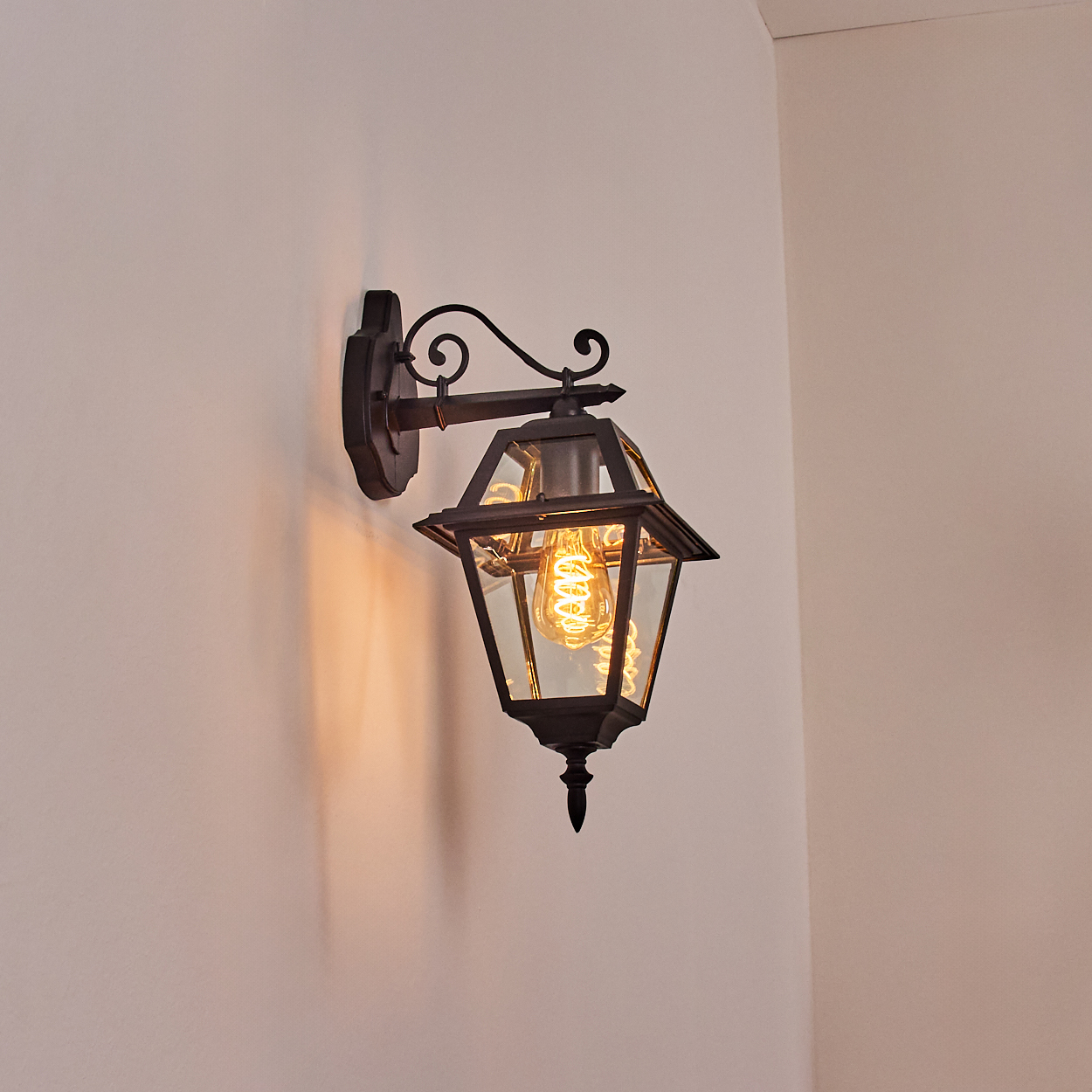 Eine schwarze Wandlaterne mit einer leuchtenden, spiralförmigen Glühbirne hängt an einer weißen Wand. Die Laterne hat Glasscheiben.