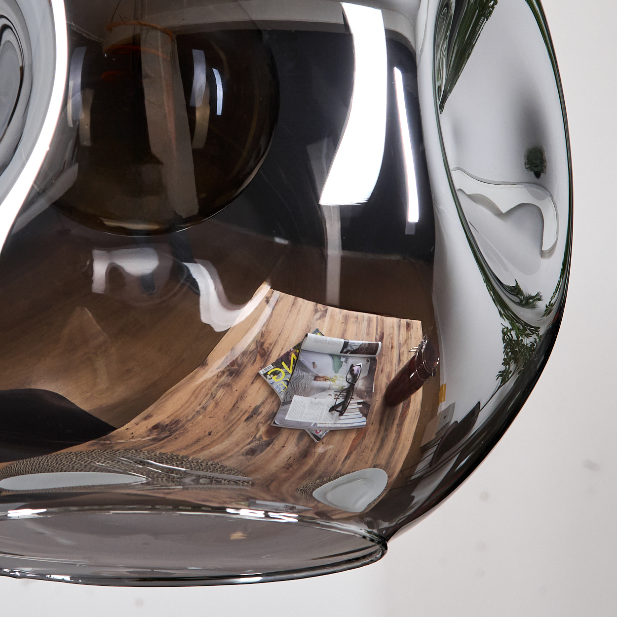 Ein großes, transparentes, gebogenes Objekt reflektiert eine Holzoberfläche mit Zeitschriften und Gläsern. Der Hintergrund ist weiß mit grünen Pflanzen.