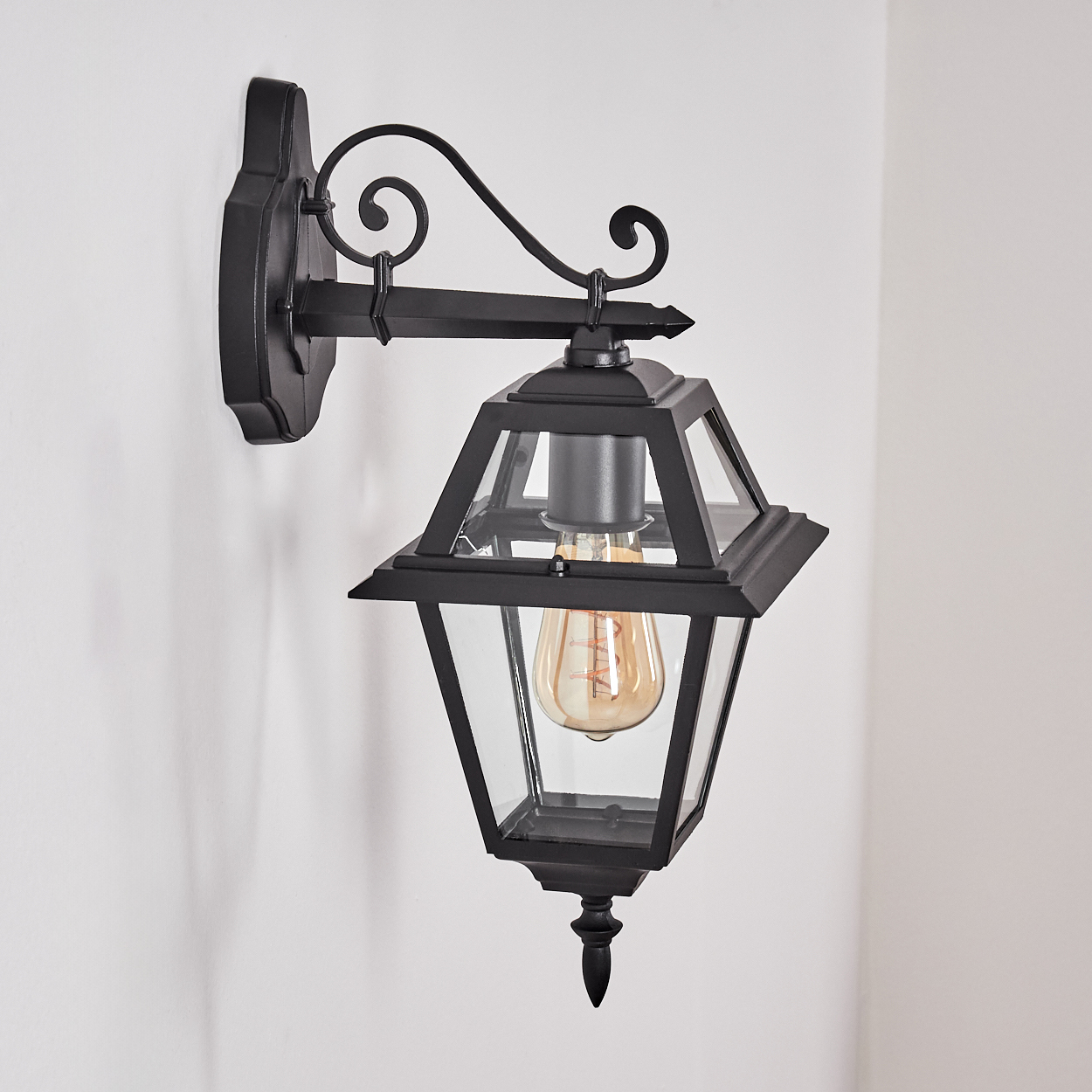 Eine schwarze Außenwandlampe mit einer Vintage-Glühbirne ist an einer weißen Wand montiert. Die Lampe hat ein Glasgehäuse.