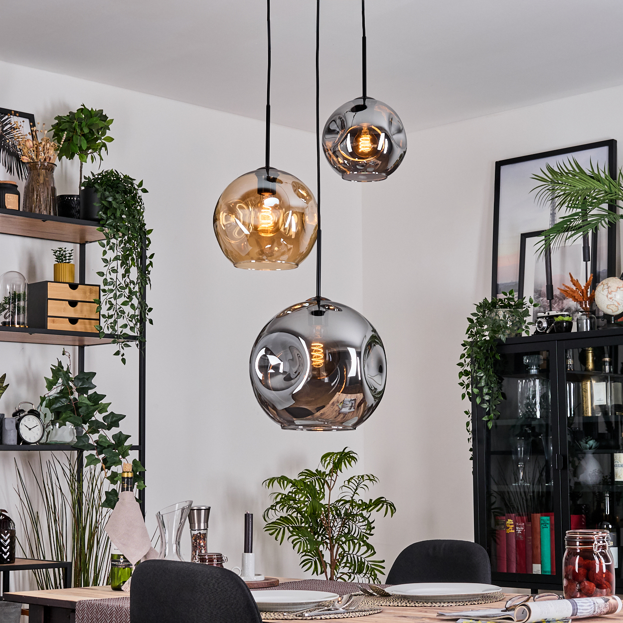 Drei Pendelleuchten mit Glaskugeln hängen über einem Esstisch. Der Raum hat ein Regal mit Pflanzen und einen Schrank mit Dekorationen.