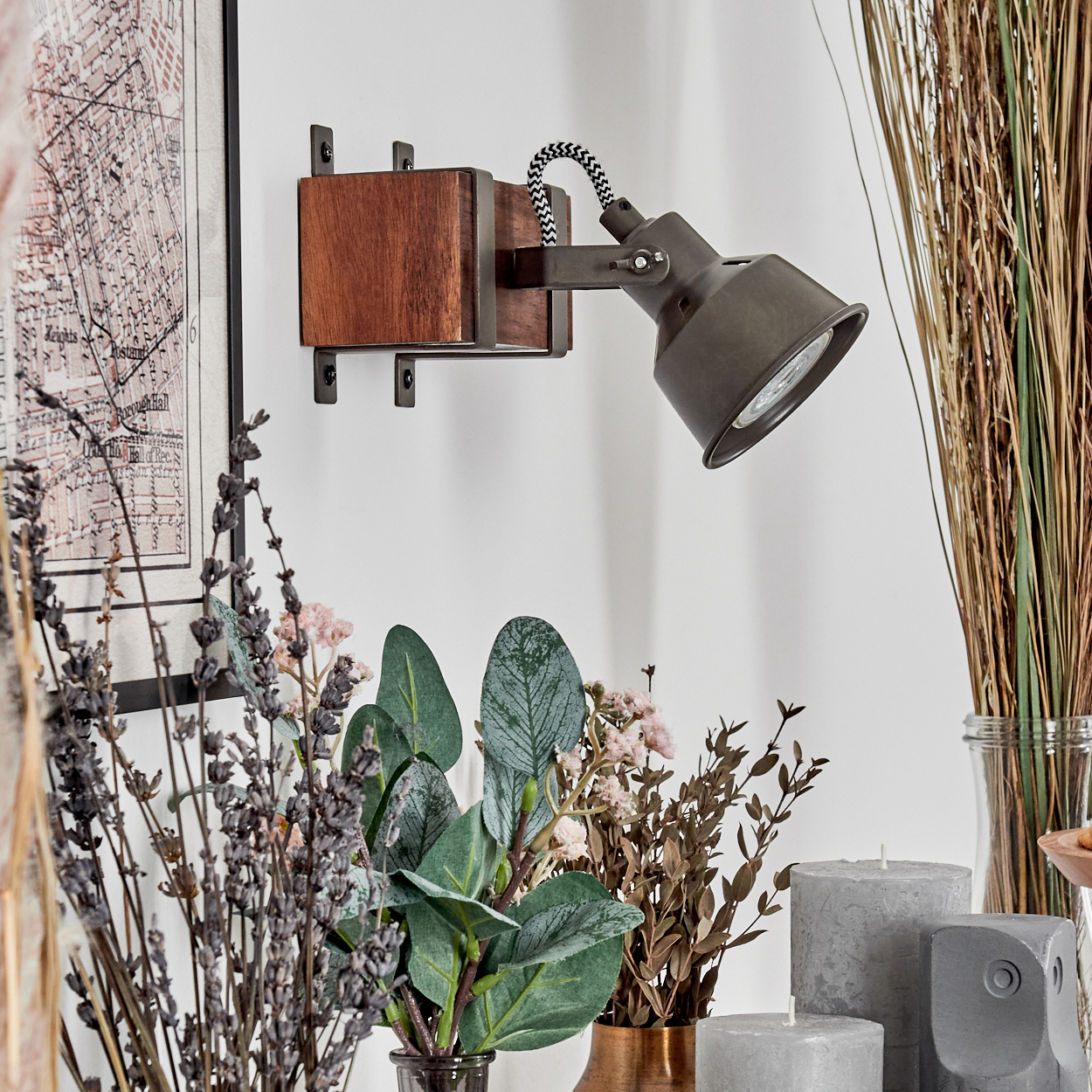 Eine dunkle Metalllampe auf einem Holzblock. Es gibt Blumen in Vasen, Kerzen und eine Karte an der Wand.
