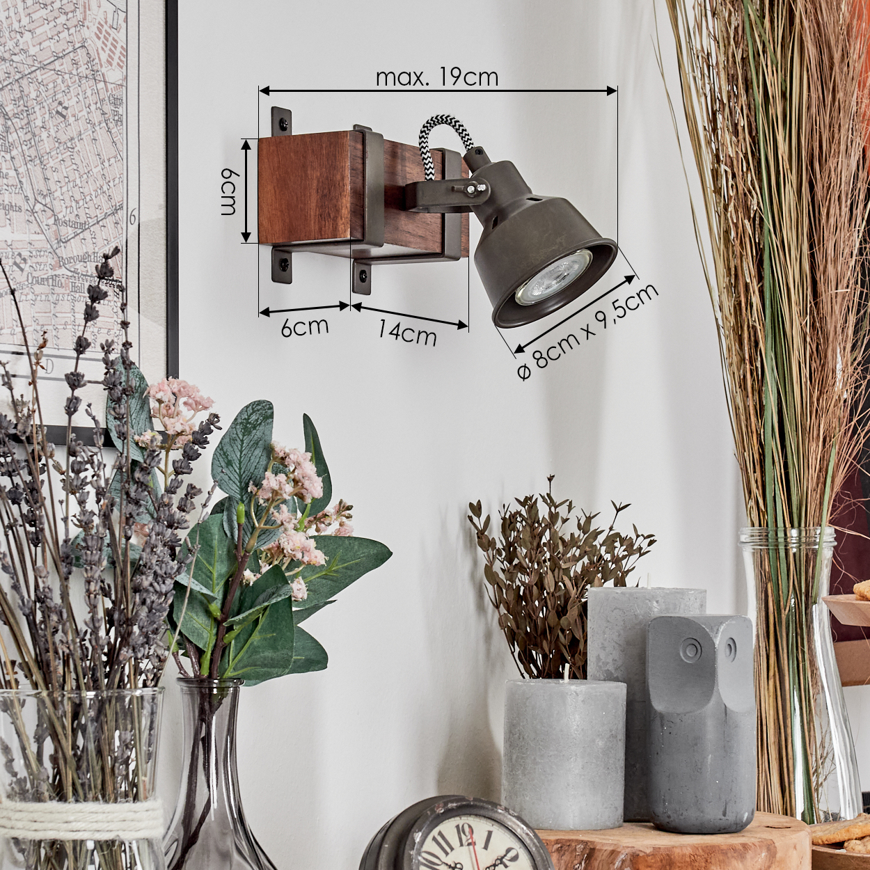 Eine Wandlampe mit Holzsockel und schwarzem Lampenkopf. Vor einer weißen Wand stehen Vasen mit Blumen und Kerzen.