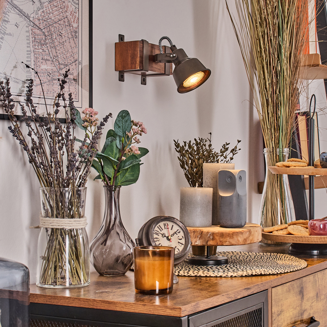Ein Raum mit Holztisch, Lampe, Blumen in Vasen, Kerzen, Uhr und Karte an der Wand. Die Szene wirkt gemütlich und warm.
