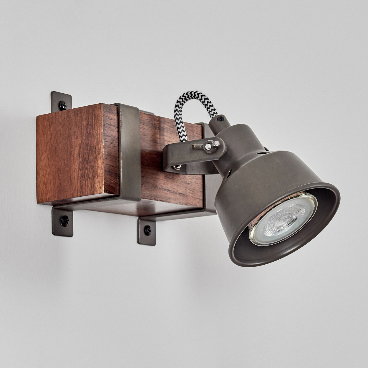 Ein an der Wand montierter Strahler. Er hat eine Holzbasis mit Metallhalterungen und einen dunkelgrauen Scheinwerfer. Das Kabel hat ein schwarz-weißes Muster.
