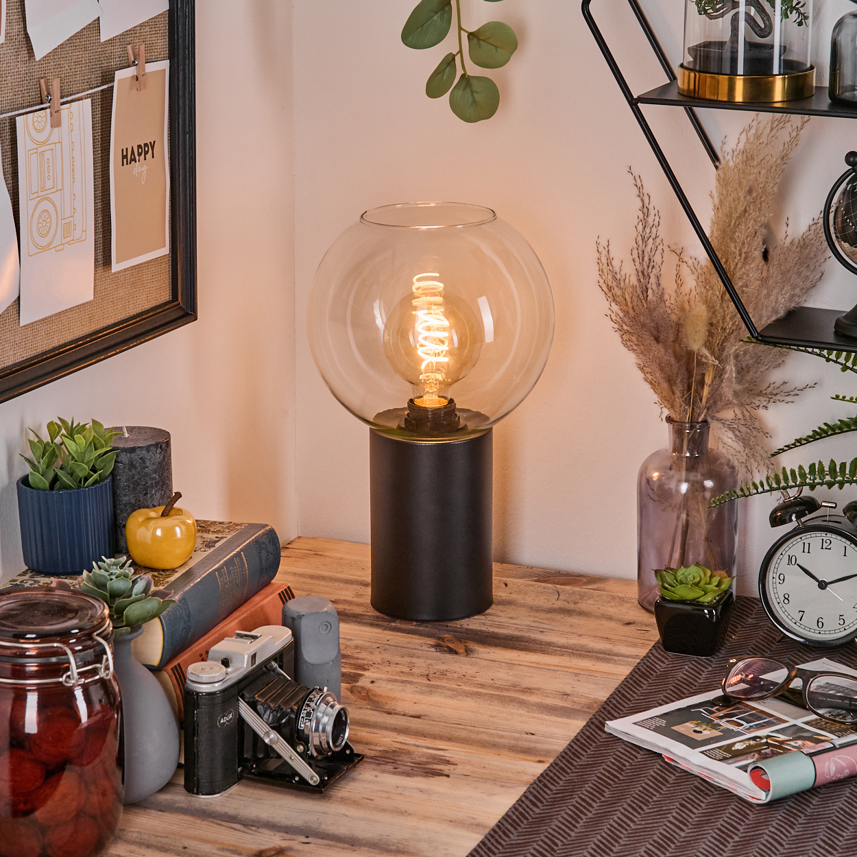 Eine beleuchtete Lampe auf einem Holztisch mit Büchern, einer Kamera und einem Glas. Im Hintergrund ist eine Pinnwand zu sehen.