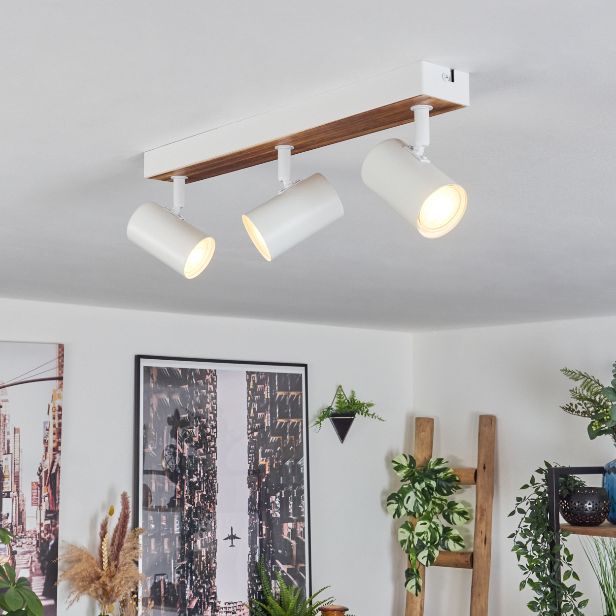 Deckenlampe mit drei weißen Strahlern. Darunter ein gerahmtes Bild und Pflanzen. Eine Holzleiter und weitere Pflanzen sind sichtbar.