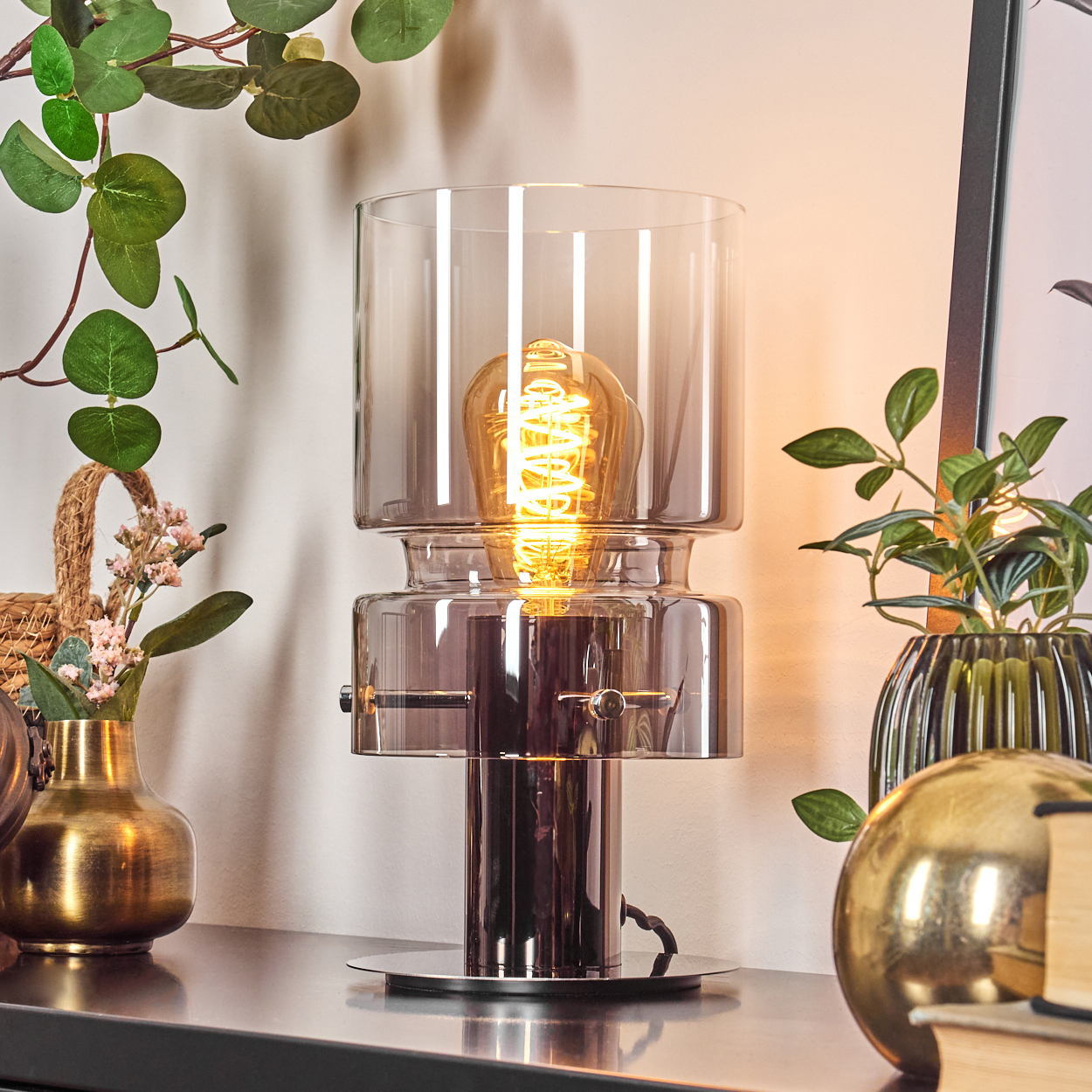 Eine moderne Lampe mit leuchtender Glühbirne steht auf einem schwarzen Tisch. Dekorative Pflanzen und eine goldene Kugel umgeben die Lampe.
