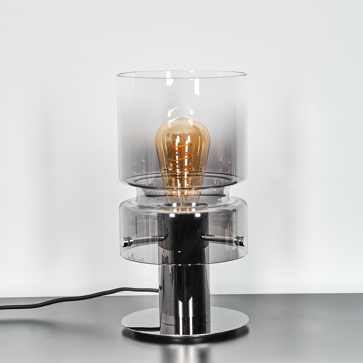 Eine moderne Lampe mit einer zentralen Glühbirne. Sie hat einen Chromfuß, einen Glasschirm und steht auf einer reflektierenden Oberfläche vor einer weißen Wand.