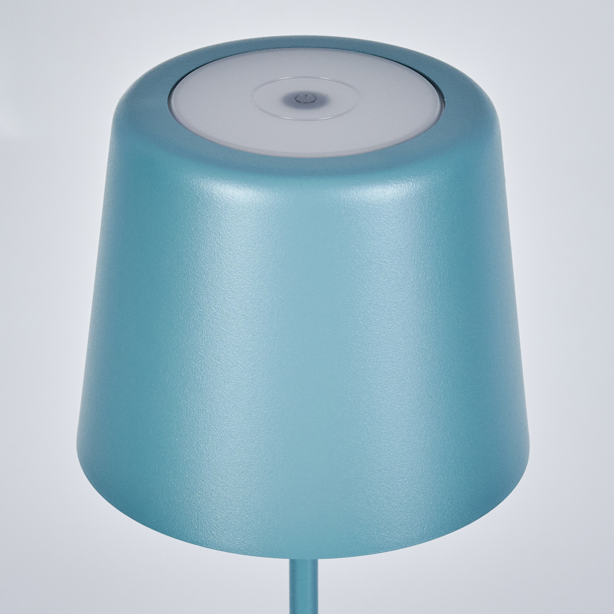 Eine türkisblaue Lampe mit weißer Oberseite. Sie hat einen runden Sockel und einen Stiel. Der Hintergrund ist weiß.