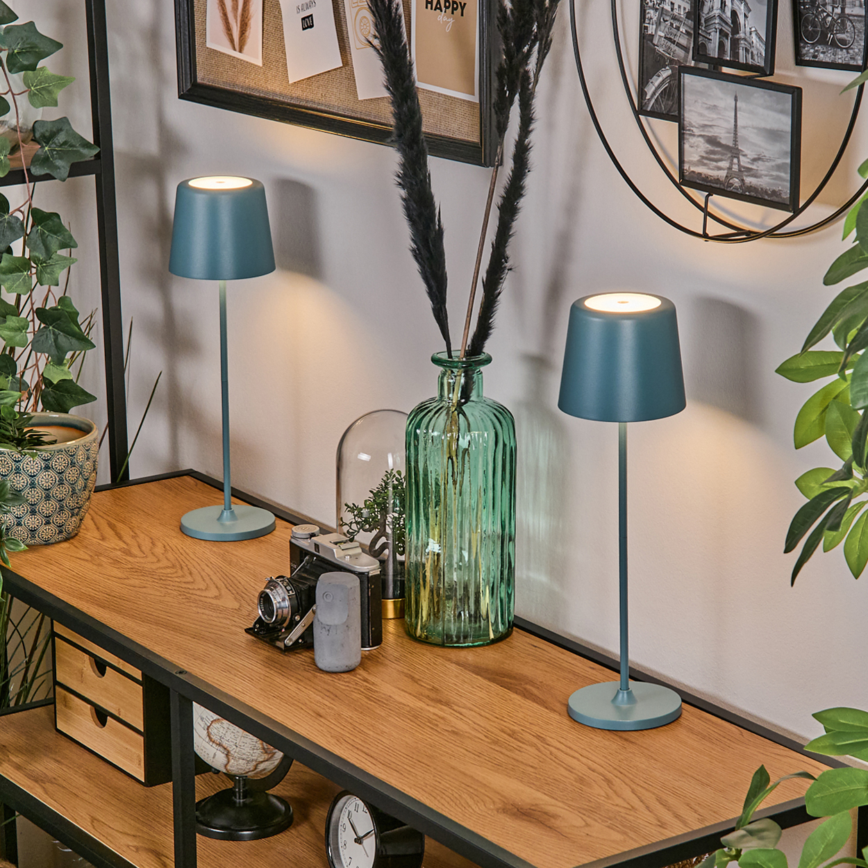 Zwei blaue Lampen auf einem Holztisch. Eine grüne Vase mit schwarzen Pflanzen, eine Kamera und ein Globus stehen ebenfalls auf dem Tisch.