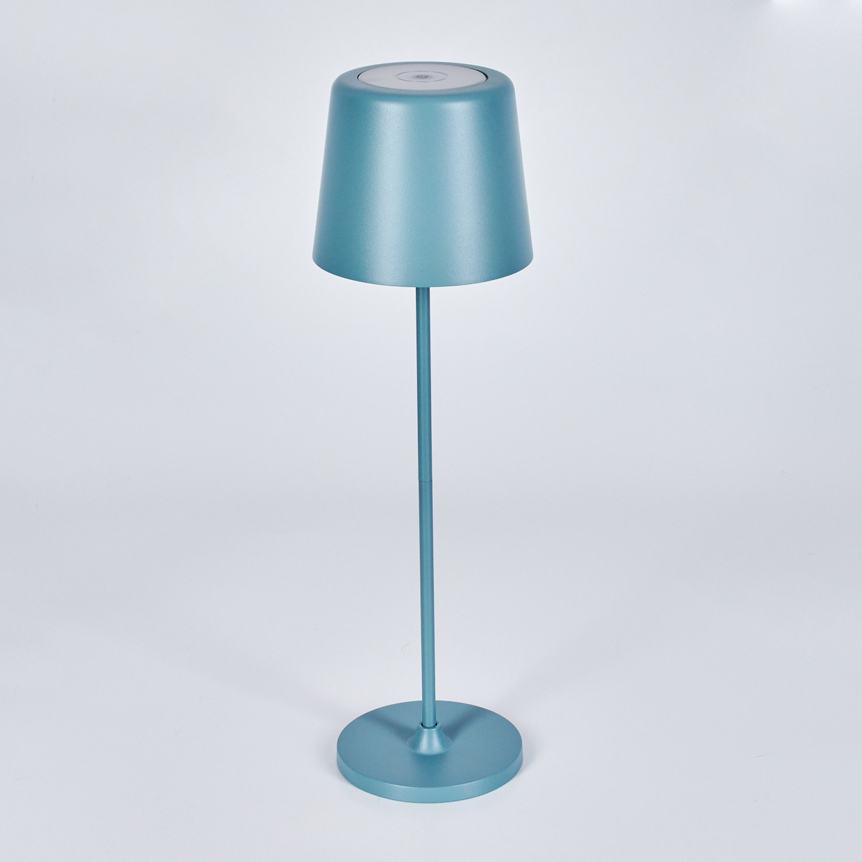 Eine türkisblaue Lampe steht auf einer weißen Oberfläche. Sie hat einen runden Fuß, einen langen Stiel und einen Lampenschirm. Der Hintergrund ist weiß.