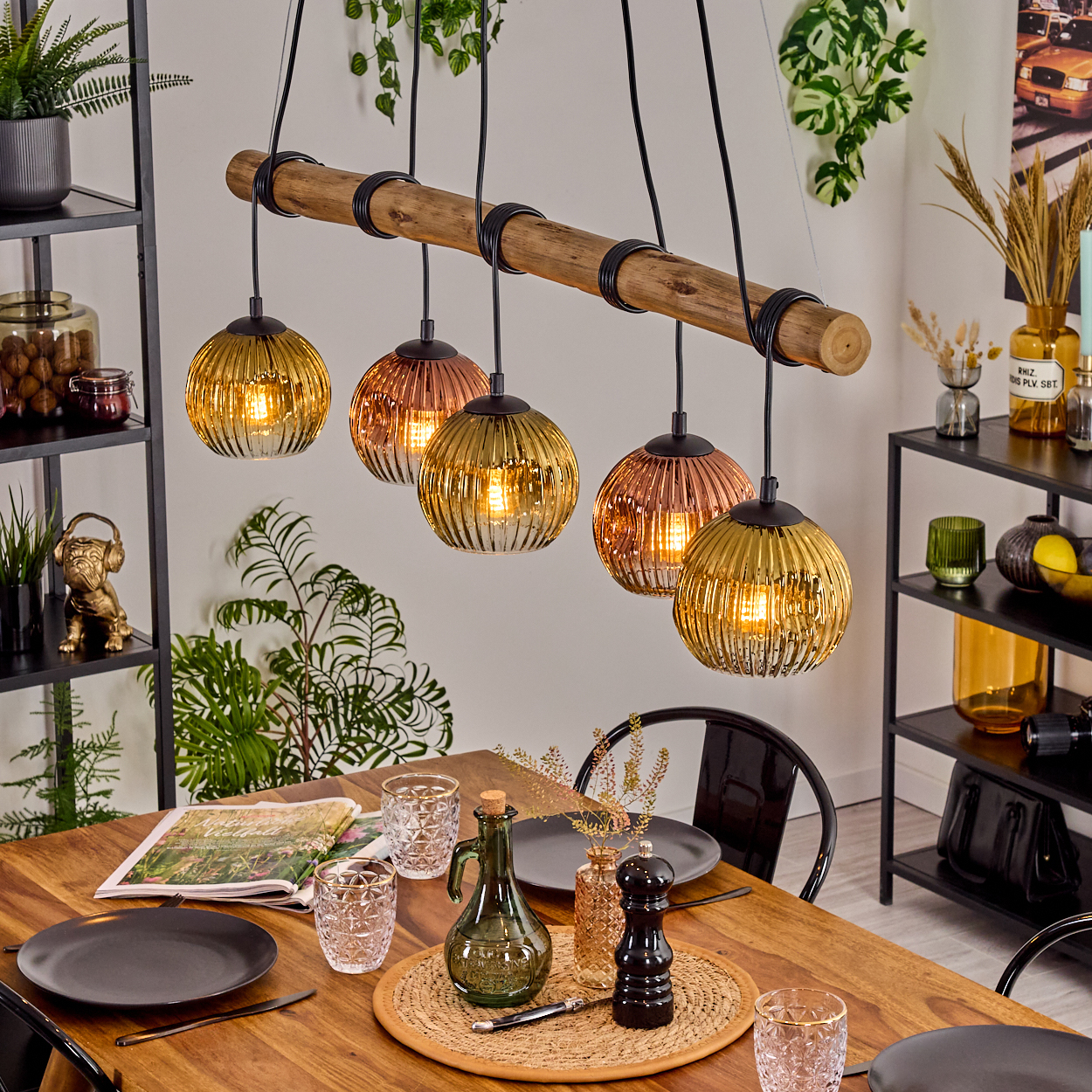 Esszimmer mit Holztisch, Glaslampen und Dekorationsartikeln. Schwarze Regale mit Pflanzen und Ornamenten sind sichtbar.