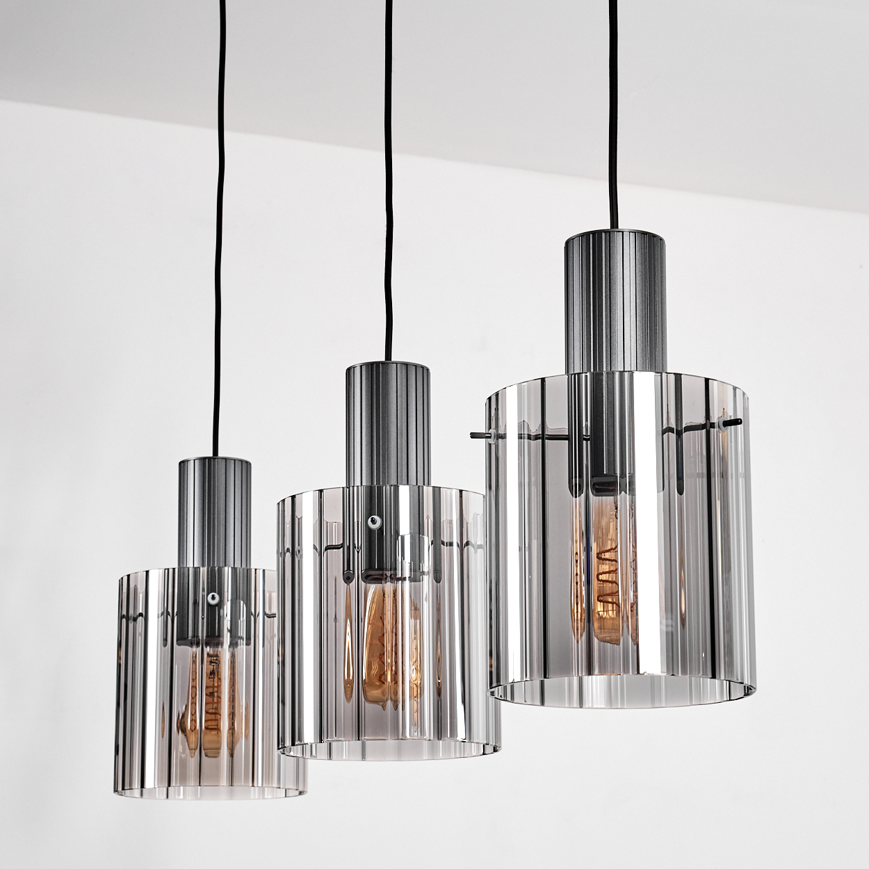 Drei Pendelleuchten mit zylindrischen Glasschirmen, Edison-Glühbirnen und schwarzen Kabeln vor weißem Hintergrund.