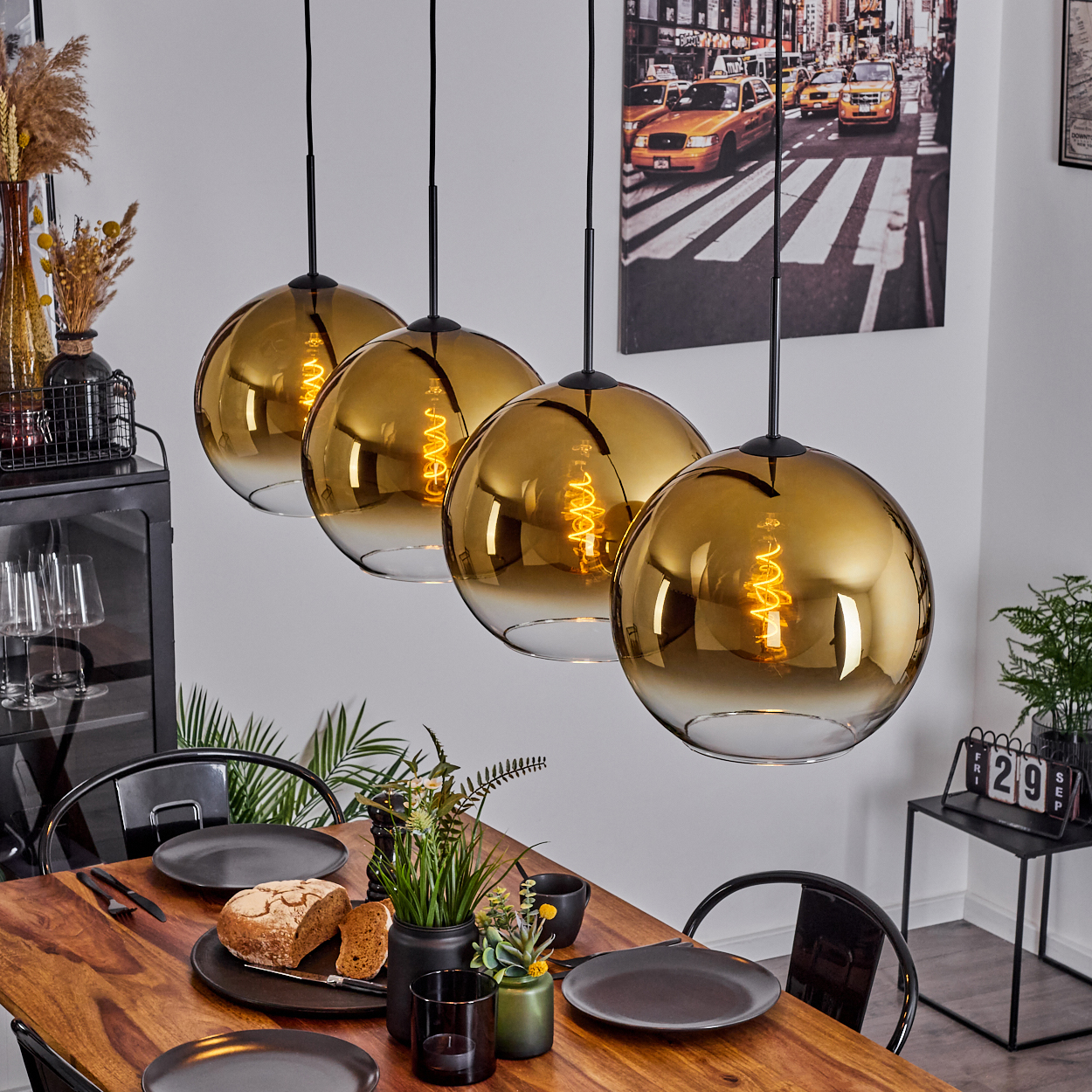 Esstisch mit vier goldenen Pendelleuchten. Brot, Teller und Pflanzen stehen auf dem Holztisch. Ein Stadtbild an der Wand.