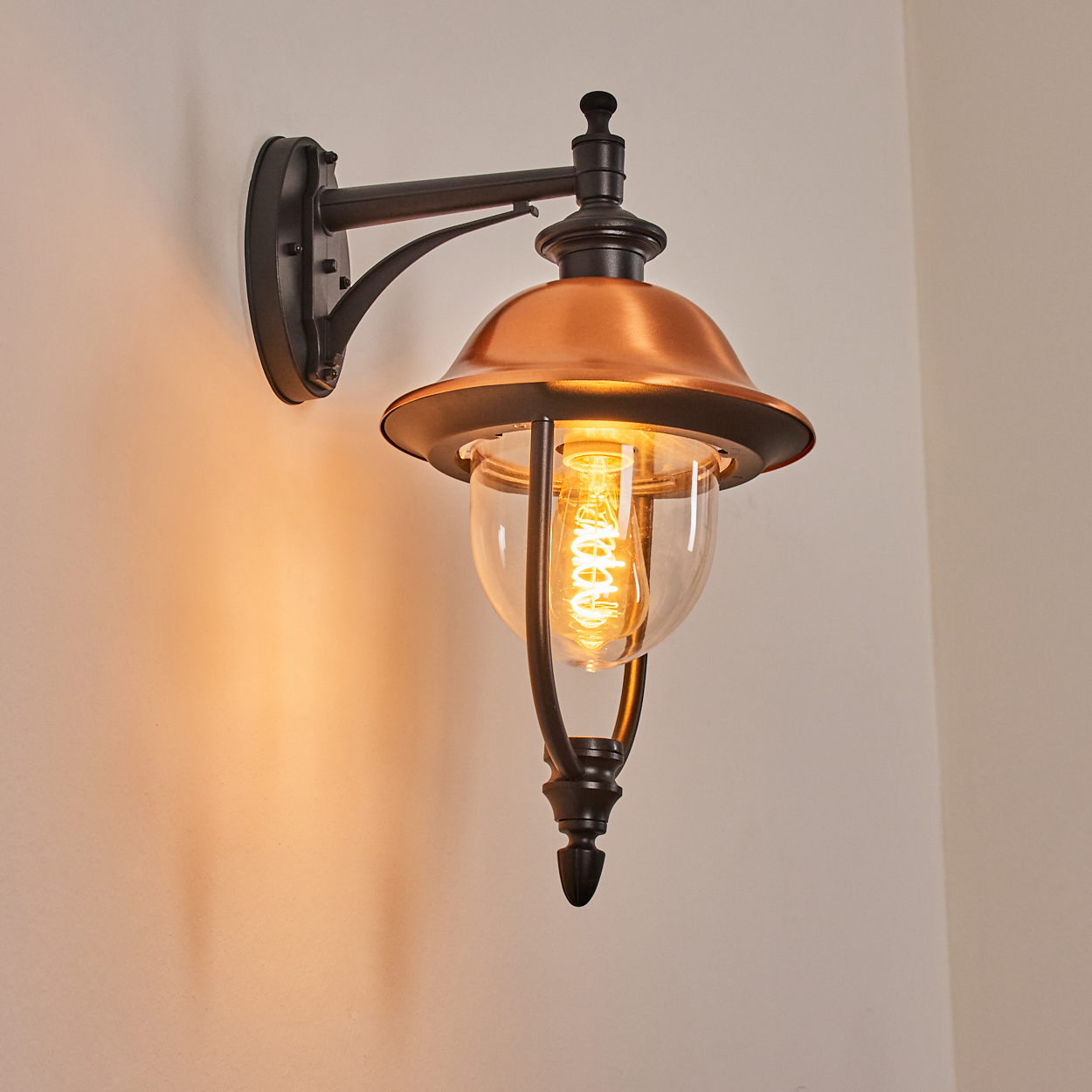 Eine an der Wand montierte Außenlampe. Sie hat eine kupferfarbene Haube und ein Glasgehäuse für die Glühbirne. Die Lampe ist schwarz.
