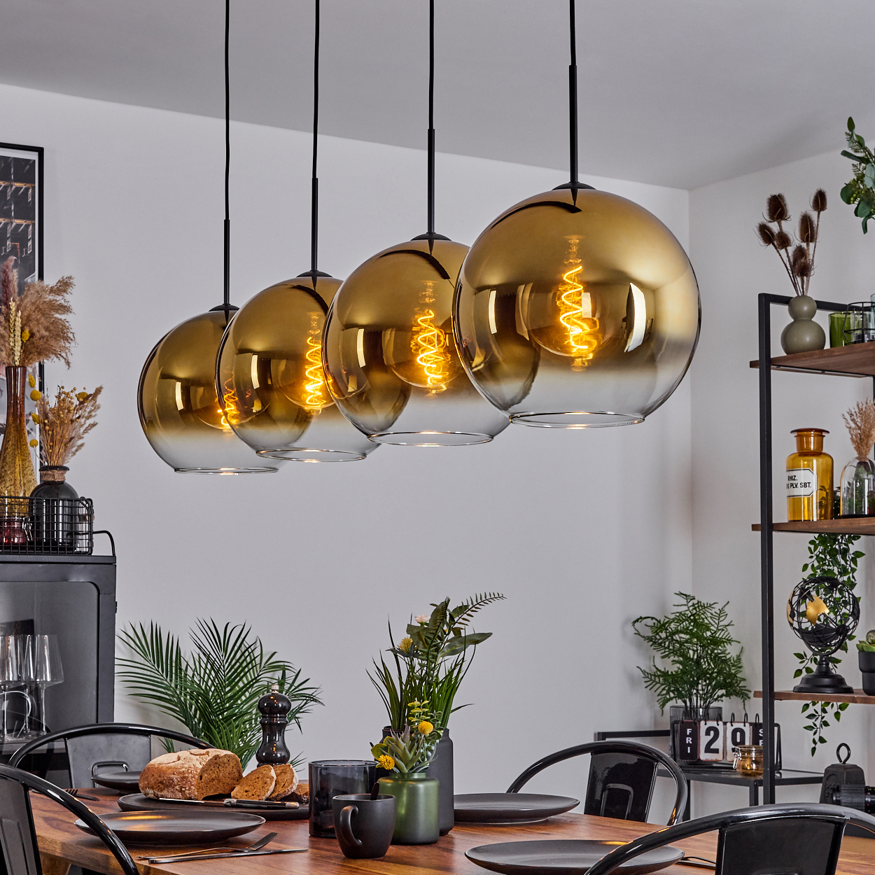 Esszimmer mit goldenen Hängelampen. Ein Tisch ist mit Brot und Tellern gedeckt. Regale mit Pflanzen im Hintergrund.