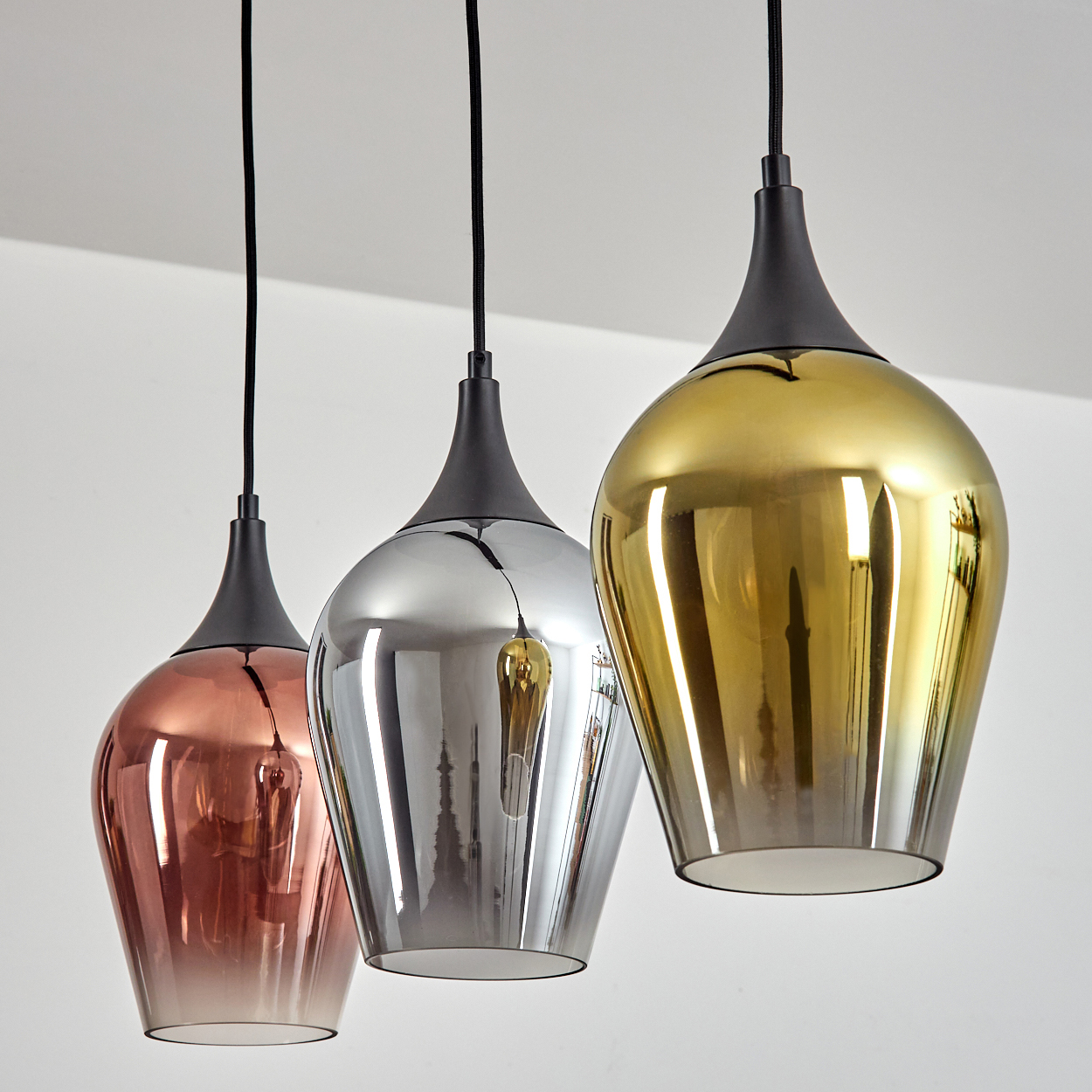 Drei Pendelleuchten mit metallischen Schirmen, Kupfer, Silber und Gold, hängen an schwarzen Kabeln vor einer weißen Decke.