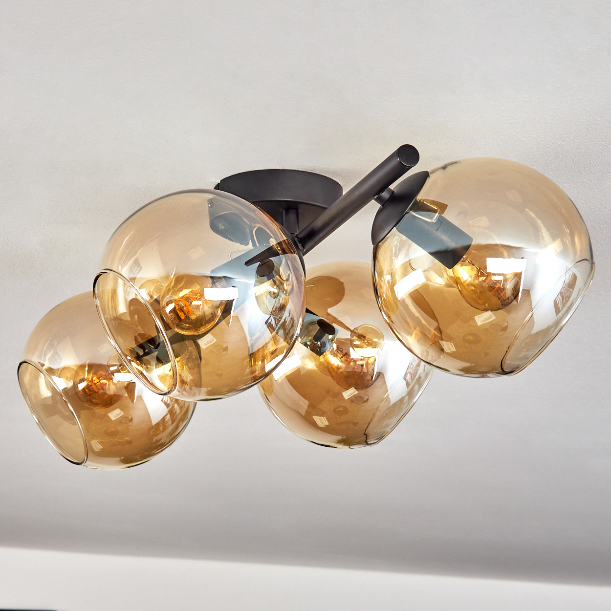 Deckenleuchte mit vier bernsteinfarbenen Glaskugeln. Schwarze Metallarme und Deckenhalterung. Warme Glühbirnen sind sichtbar.