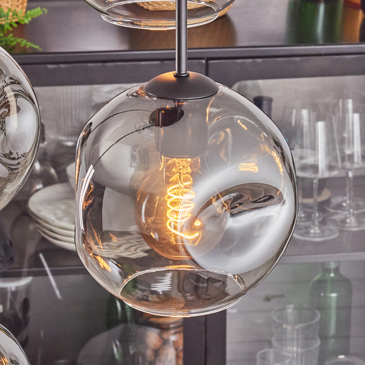 Nahaufnahme einer Glas-Pendelleuchte mit Glühbirne. Im Hintergrund Gläser und ein Schrank.