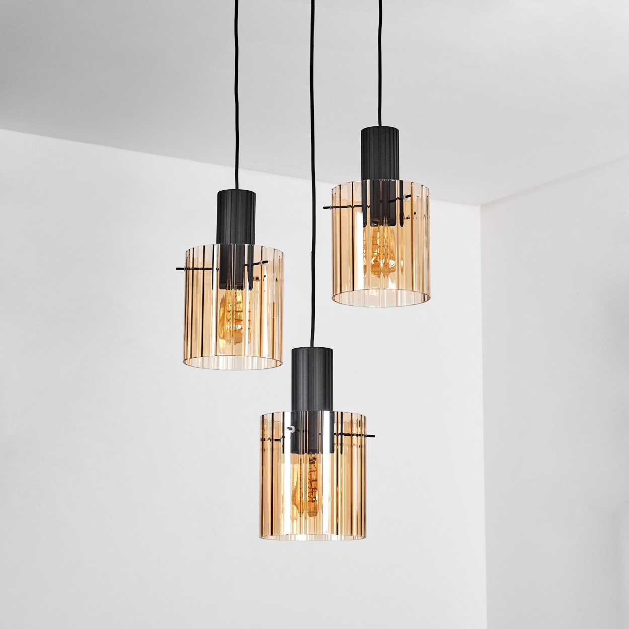 Drei Pendelleuchten mit bernsteinfarbenen Glasschirmen und schwarzen Armaturen hängen vor einer weißen Wand. Die Glühbirnen sind sichtbar.