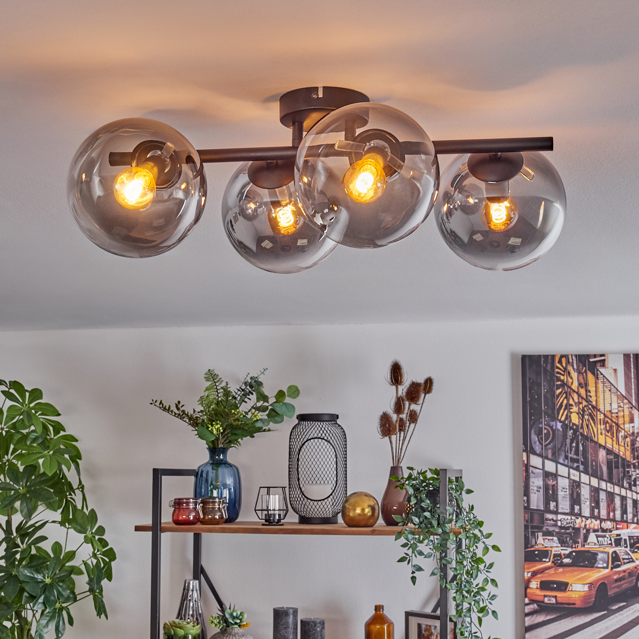 Eine Deckenleuchte mit vier Glaskugeln. Darunter ein Regal mit Vasen, Kerzen und Pflanzen. Ein NYC-Straßenkunstdruck an der Wand.