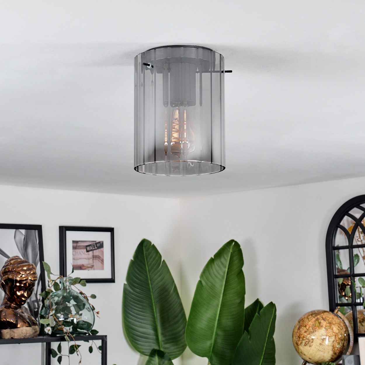 Eine moderne Deckenleuchte mit Glasschirm hängt über Pflanzen und Dekoration. Die Leuchte hat eine sichtbare Glühbirne. An der Wand hängen gerahmte Bilder.