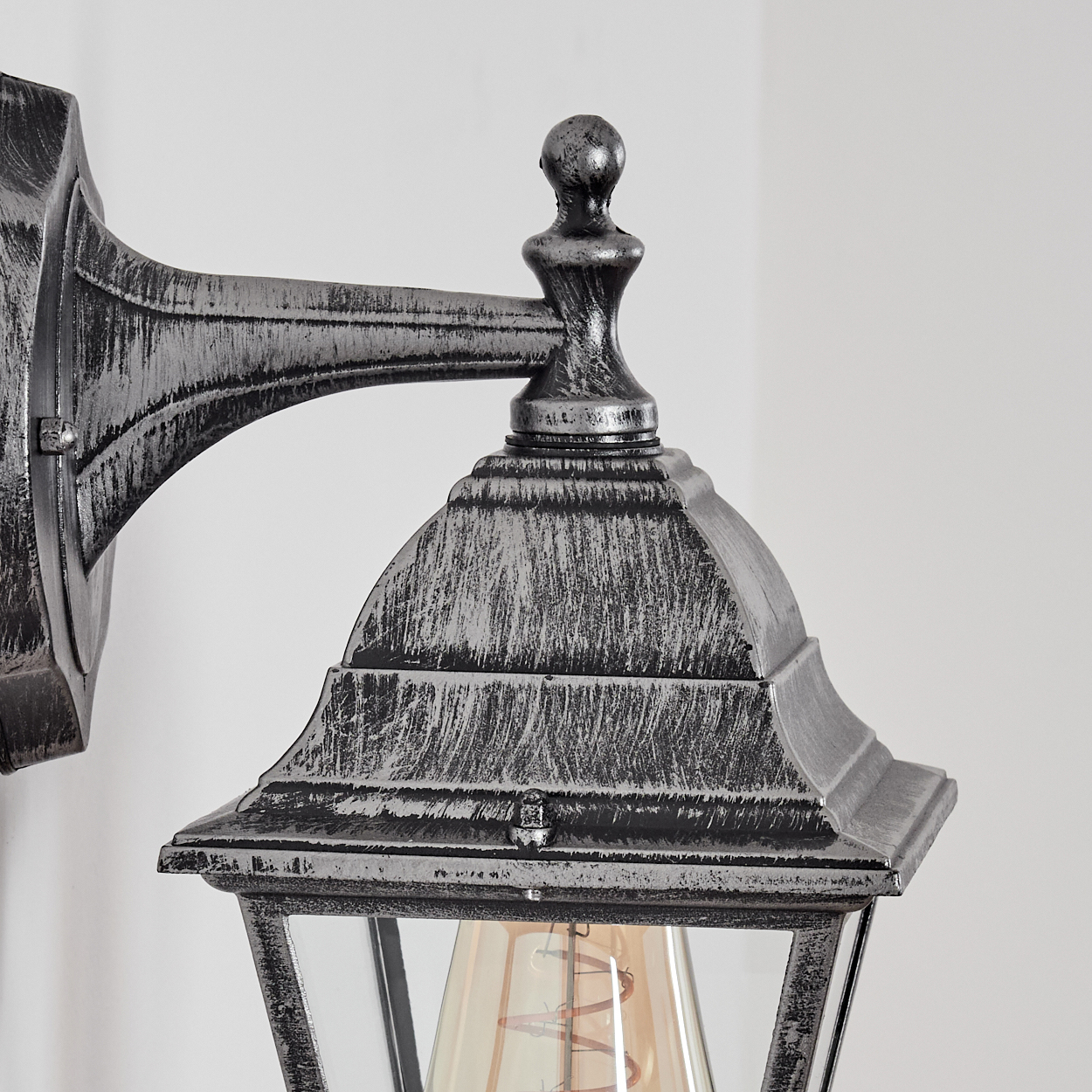 Nahaufnahme einer silbernen Außenlampe. Sie hat eine dekorative Oberseite, Glasscheiben und eine sichtbare Glühbirne. Der Hintergrund ist eine weiße Wand.