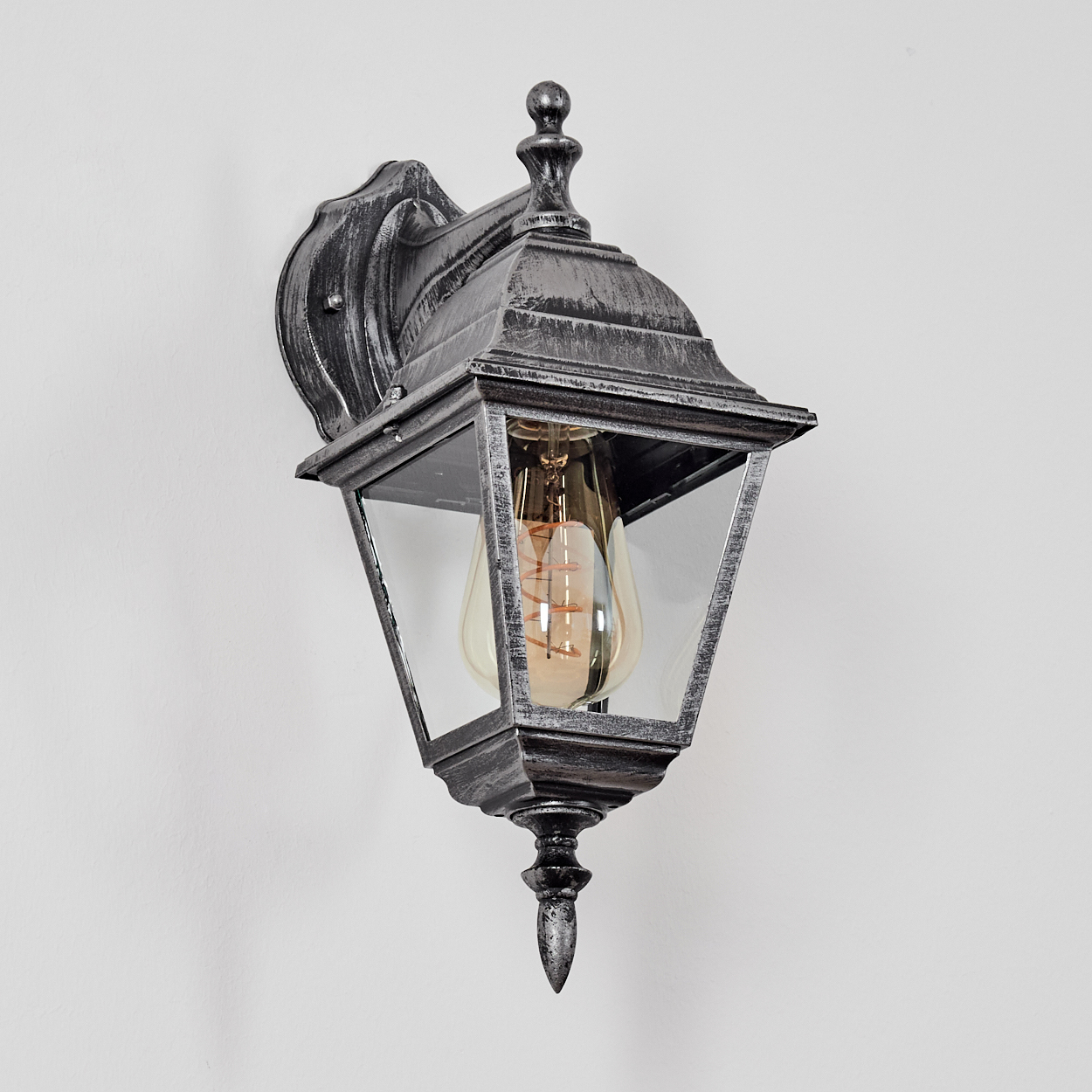 Eine metallische Wandlampe mit Glasgehäuse. Die Lampe hat ein Vintage-Design. Der Hintergrund ist eine schlichte weiße Wand.